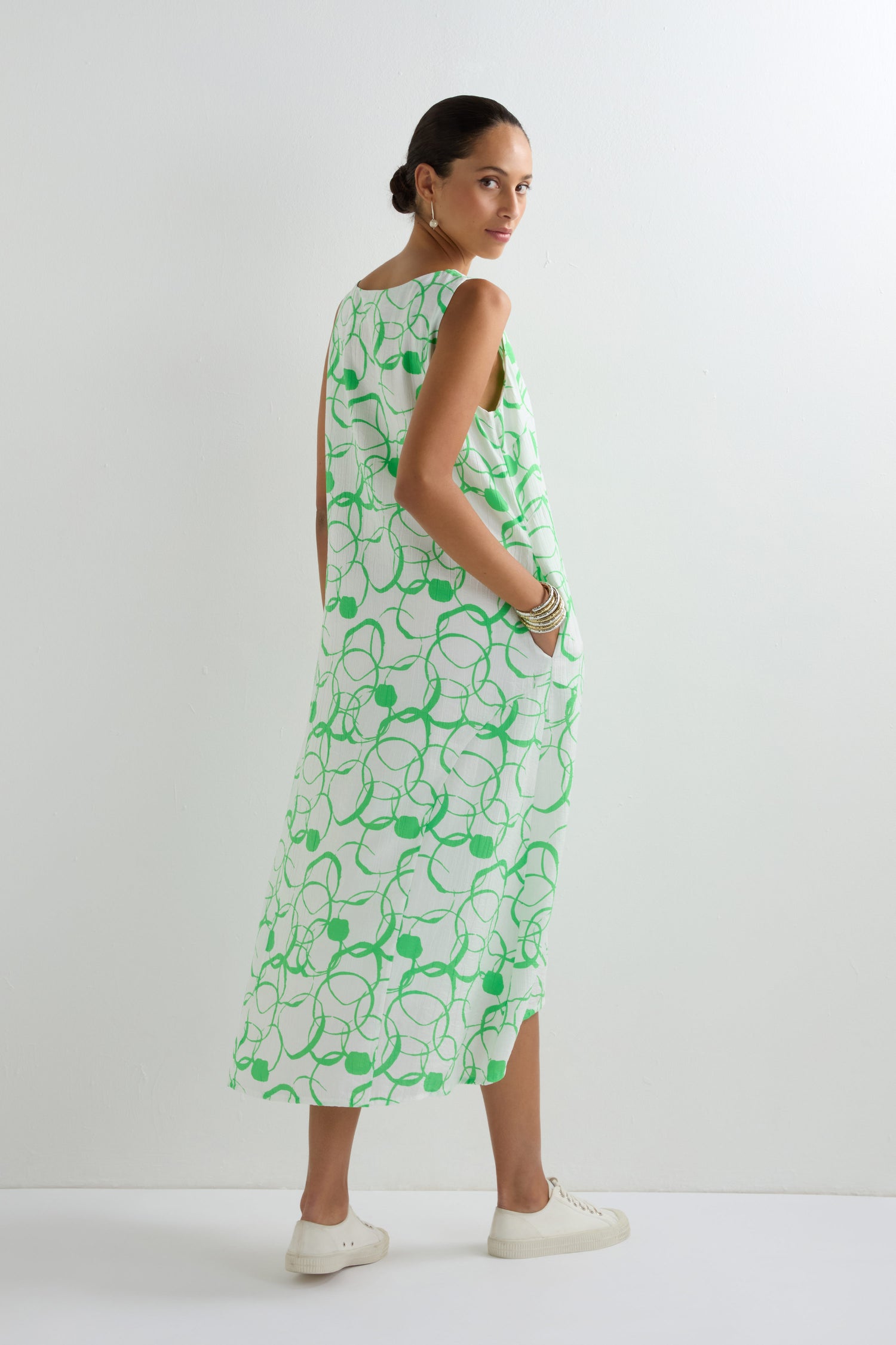 A woman stands in a white room wearing the Swirl Print Bubble Dress, which has a sleeveless bubble silhouette with a green swirl pattern. She pairs it with white sneakers and simple jewelry, looking over her shoulder.