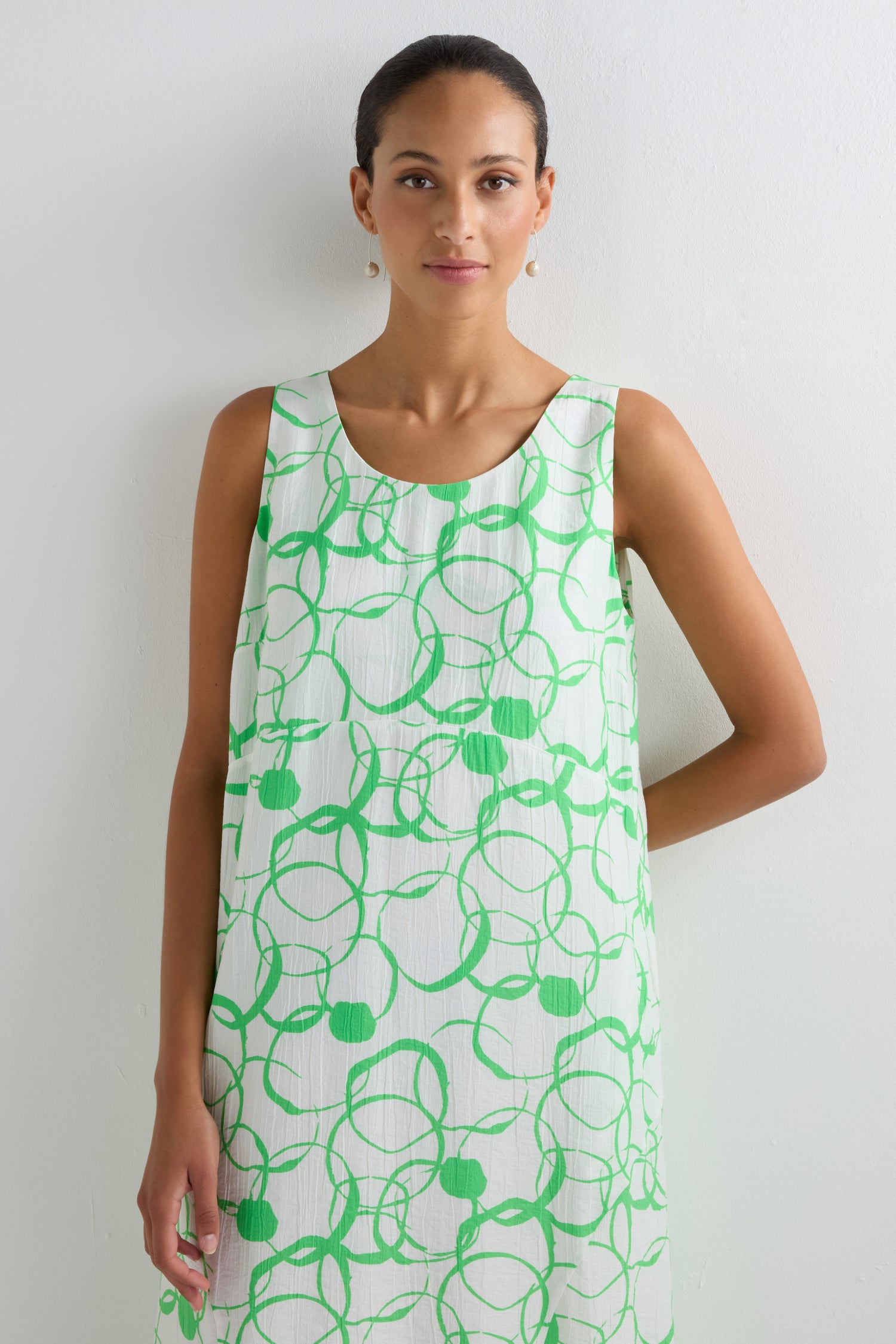 A woman stands against a white wall wearing a sleeveless Swirl Print Bubble Dress with a green swirl pattern.