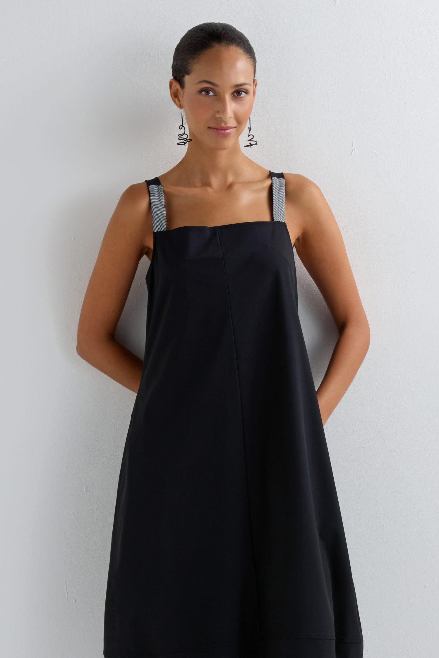 A woman stands against a white wall wearing the Stripe Strap Bubble Dress, a modern minimalist style with gray straps and geometric earrings, hands behind her back.