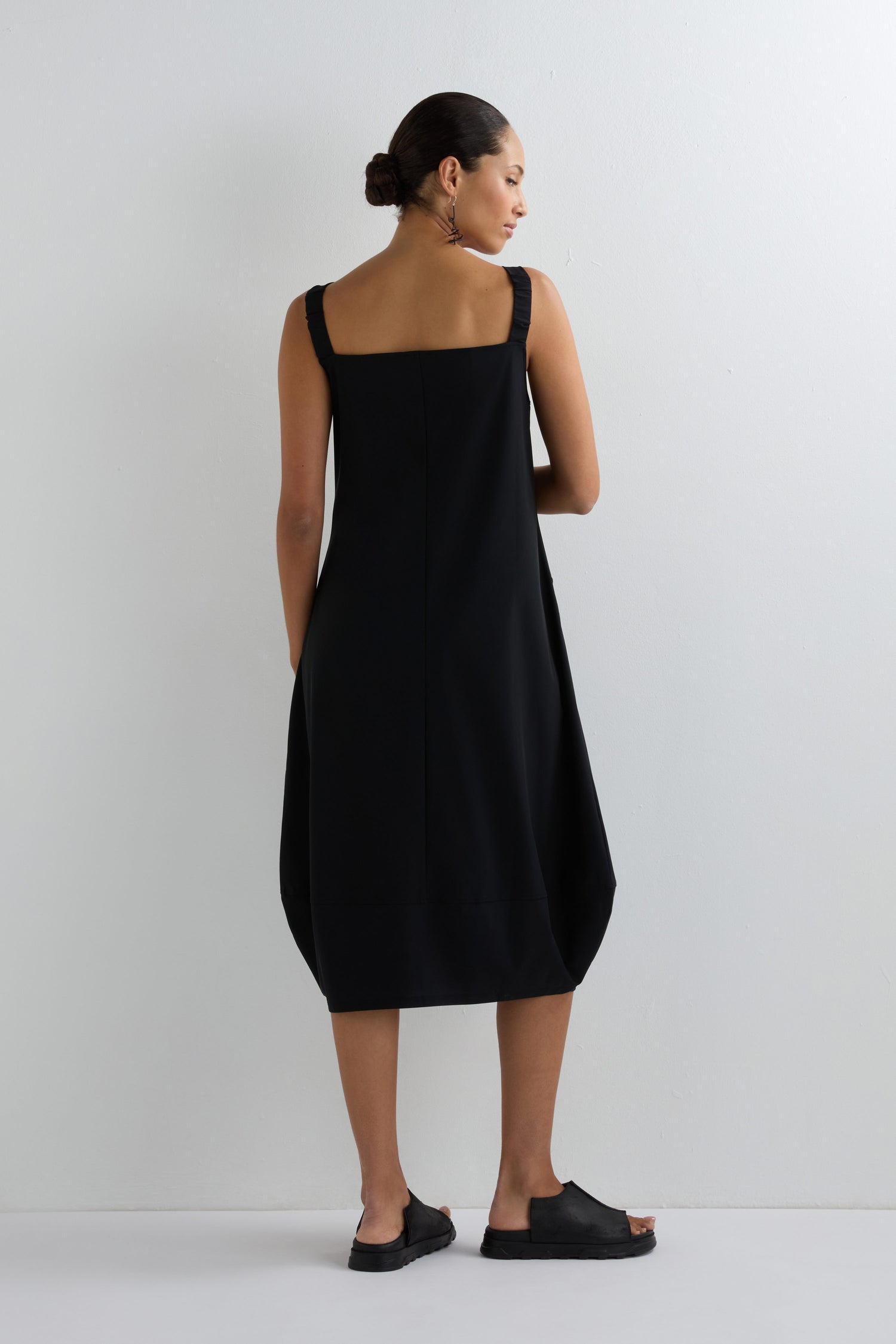 Facing a white wall, a woman wears the Stripe Strap Bubble Dress—a modern minimalist dress with wide straps and a bubble silhouette. She pairs it with black slip-on shoes and her hair is styled in a low bun.