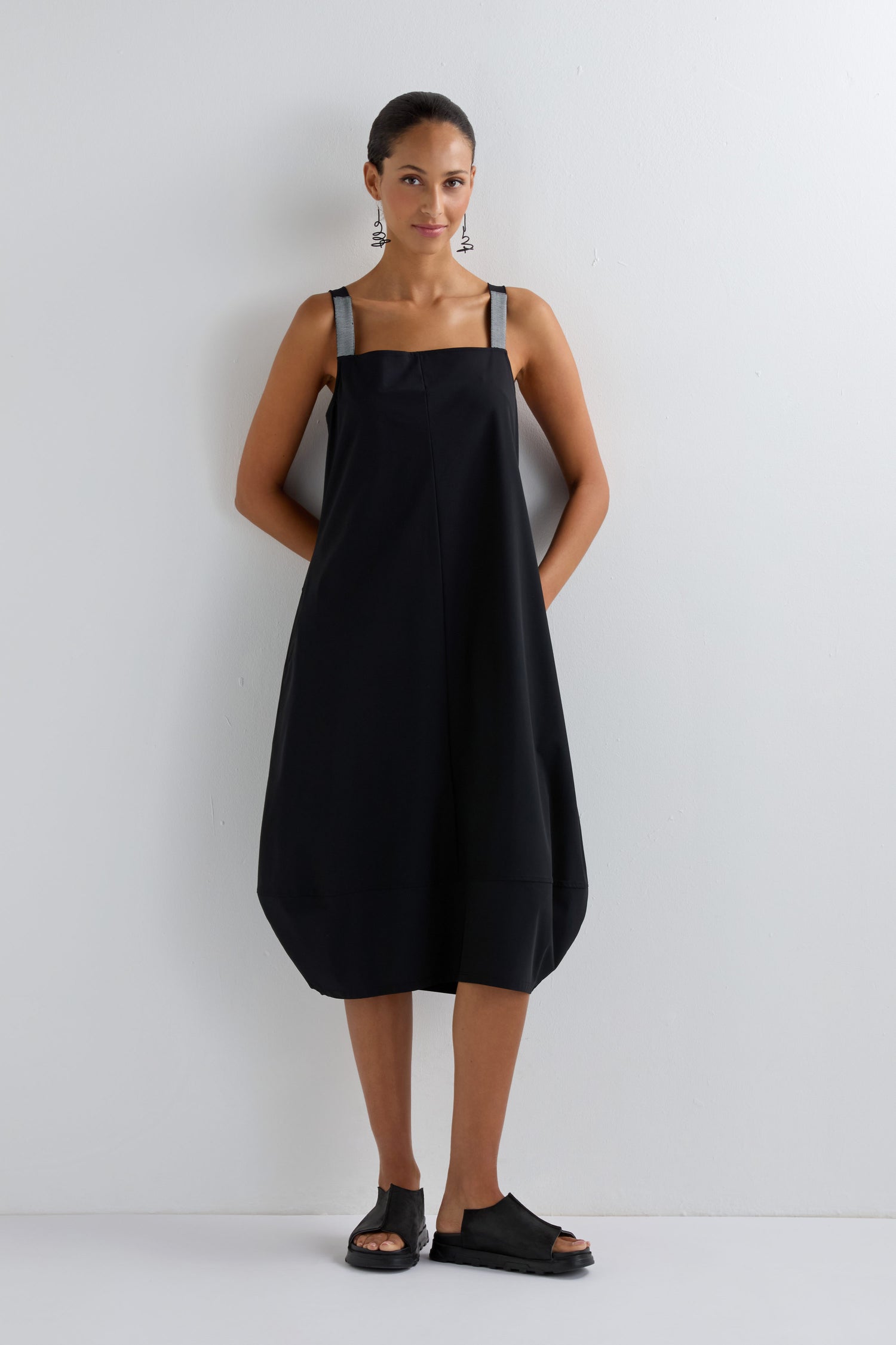 A woman stands against a white wall wearing the Stripe Strap Bubble Dress, a modern minimalist dress with thick shoulder straps, paired with black earrings and black open-toe sandals.
