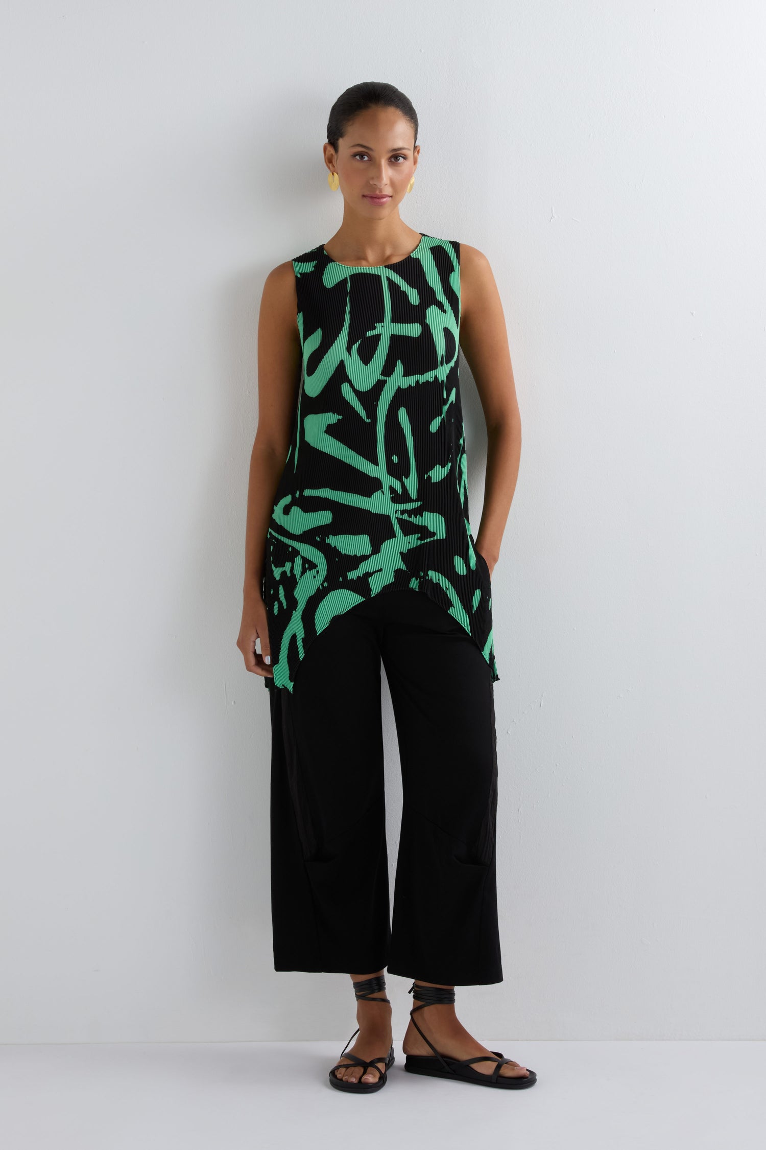 A woman stands against a white wall wearing the Long Print Pleated Vest in green and black abstract print, paired with black wide-leg pants and black sandals—a modern wardrobe essential.