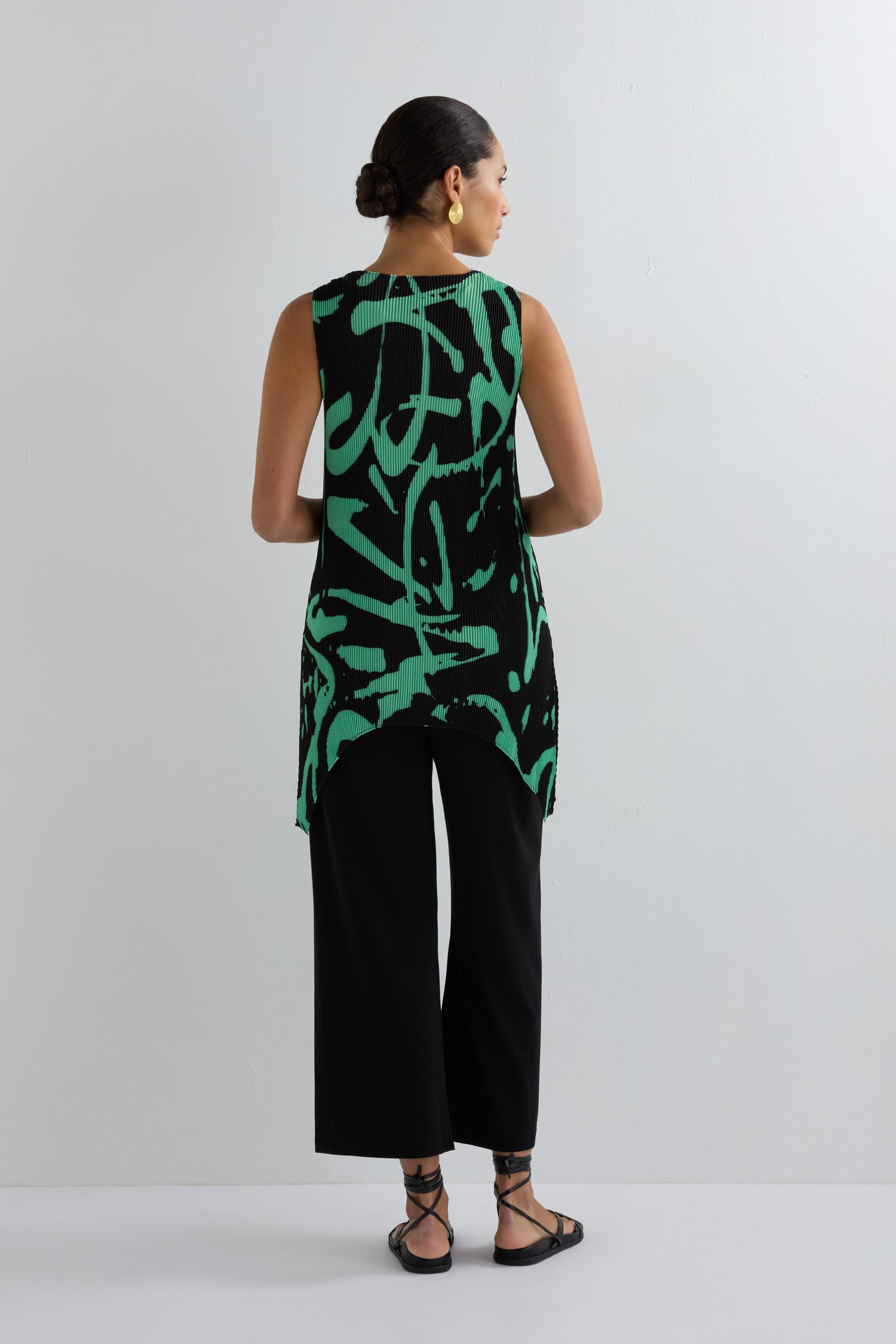 A person faces away, wearing the Long Print Pleated Vest—a sleeveless black top with green abstract patterns—paired with black wide-leg pants and sandals, set against a plain white background.