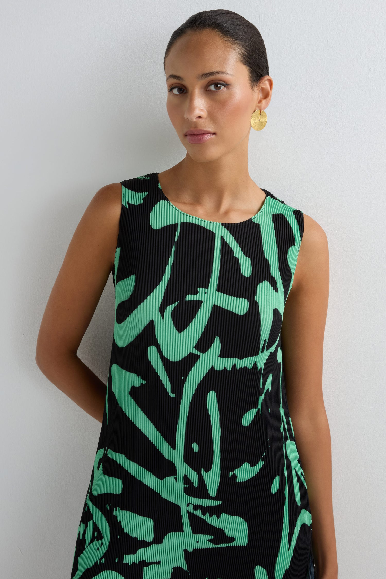 Woman wearing the Long Print Pleated Vest—a sleeveless black dress with bold green abstract design—paired with gold circular earrings and hair pulled back, standing against a white wall. A modern statement piece for any wardrobe.