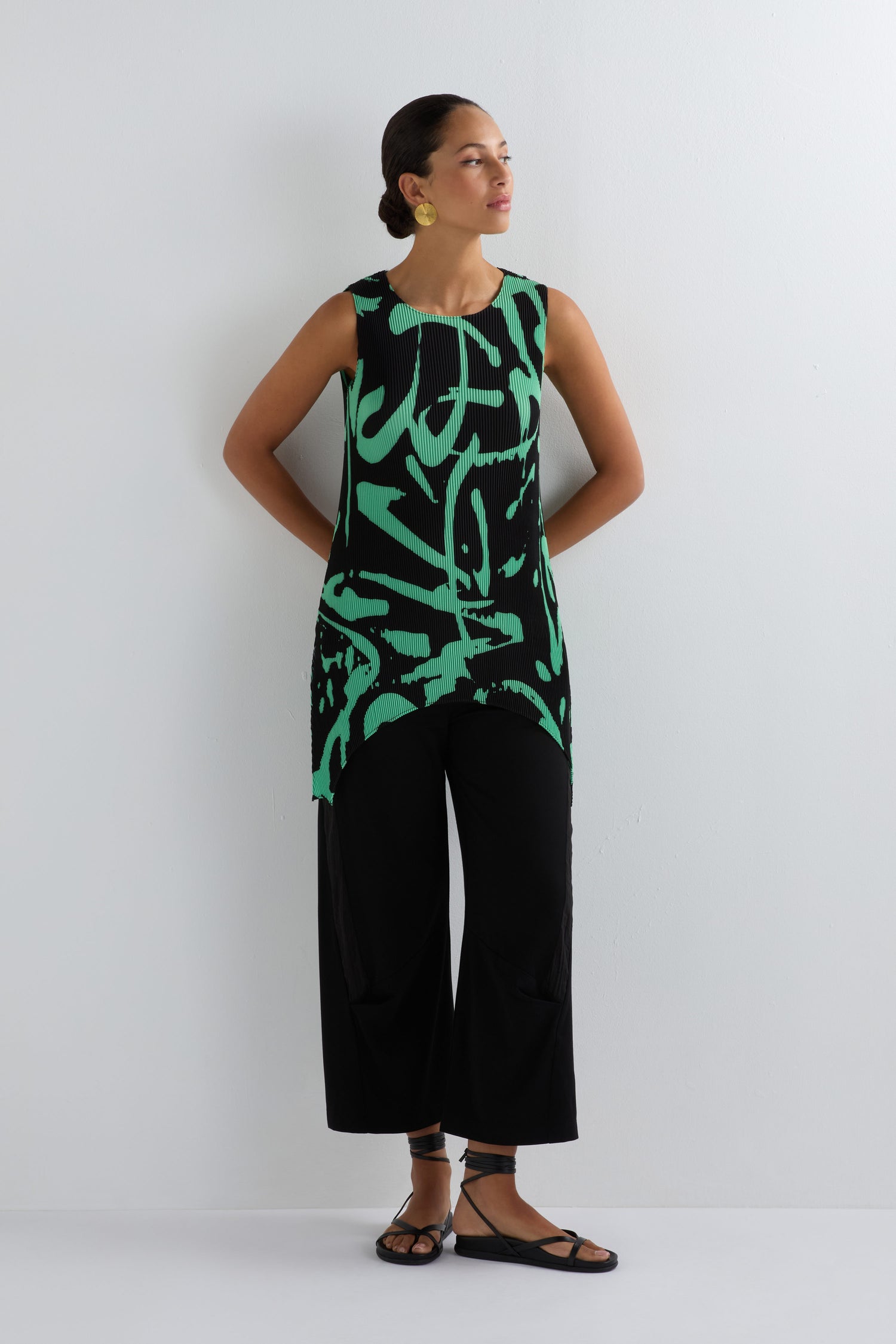 Woman stands against a white wall wearing the Long Print Pleated Vest—a black sleeveless top with a green abstract print—paired with black wide-leg pants, sandals, and gold earrings for effortless modern style.