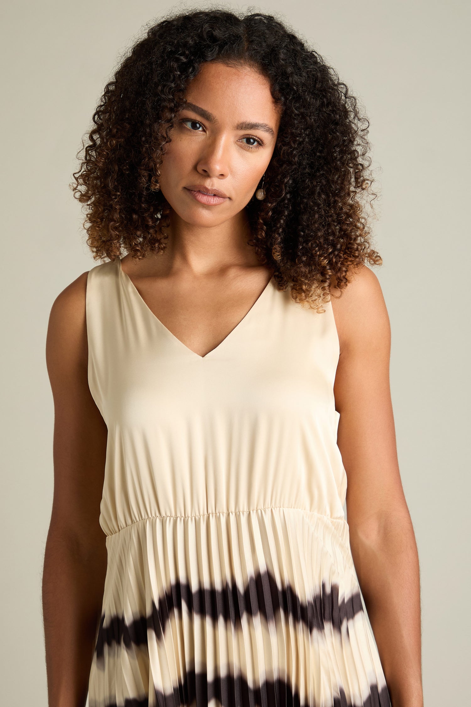 A person with curly hair wearing a Tie Dye Pleat Dress With Jersey Top, featuring a sleeveless design and gathered waist, stands in front of a neutral background.