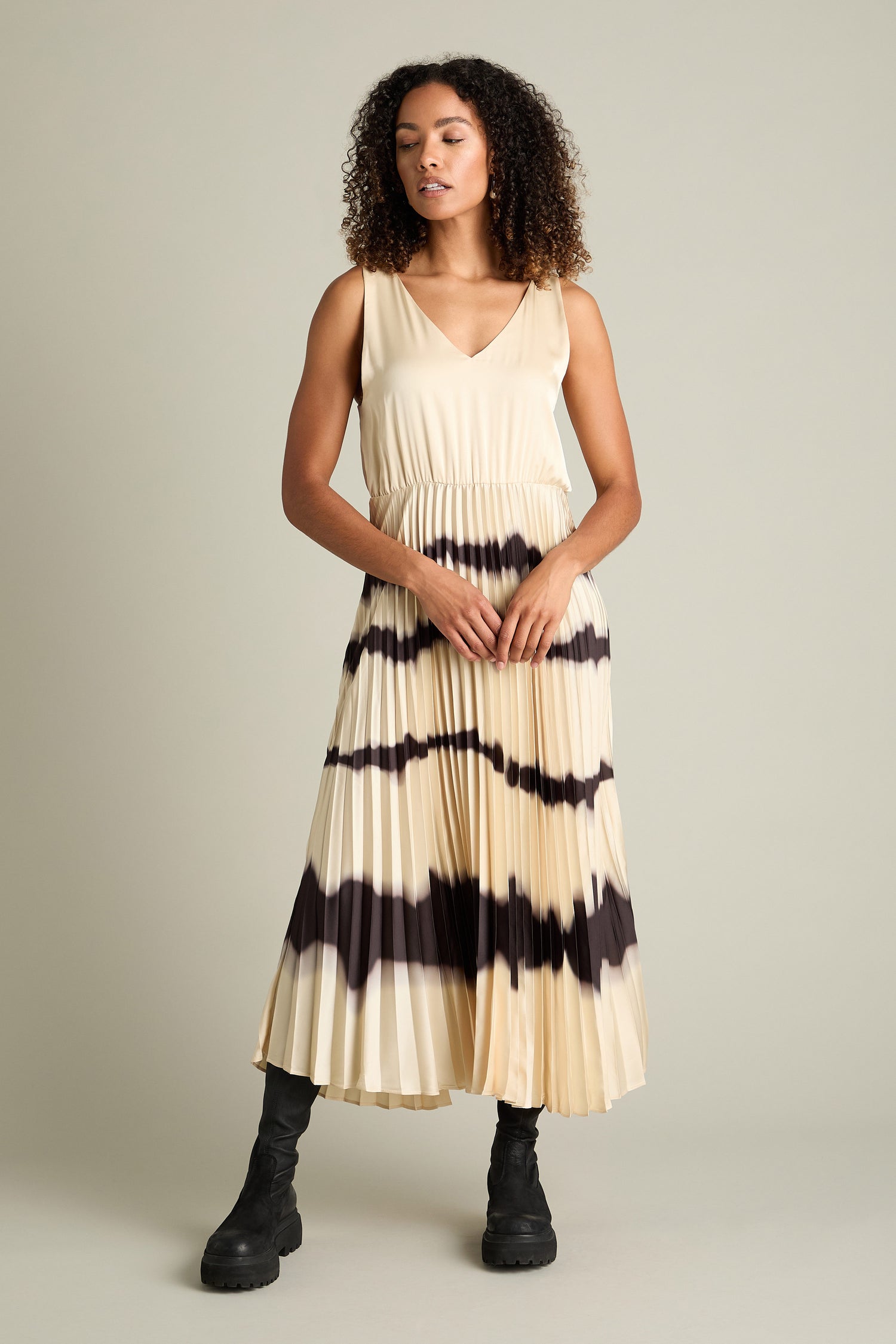 A person with curly hair wears the "Tie Dye Pleat Dress With Jersey Top" in a sleeveless beige style featuring black horizontal stripes, paired with black boots, standing against a plain background.