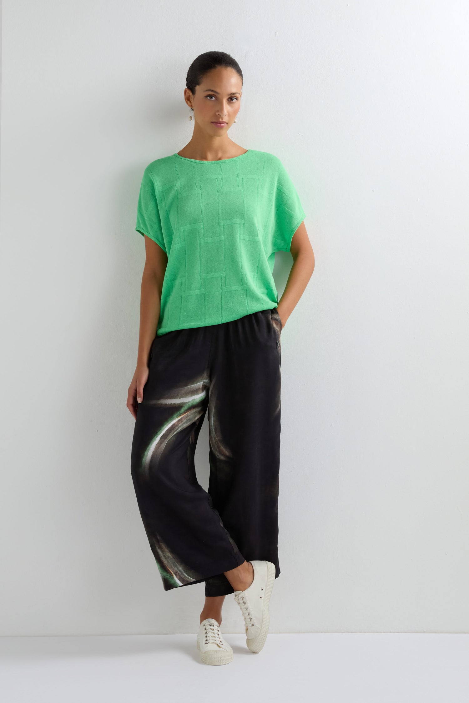 A woman stands against a white wall wearing a loose green top, Aurora Print Straight Trousers, and white sneakers.