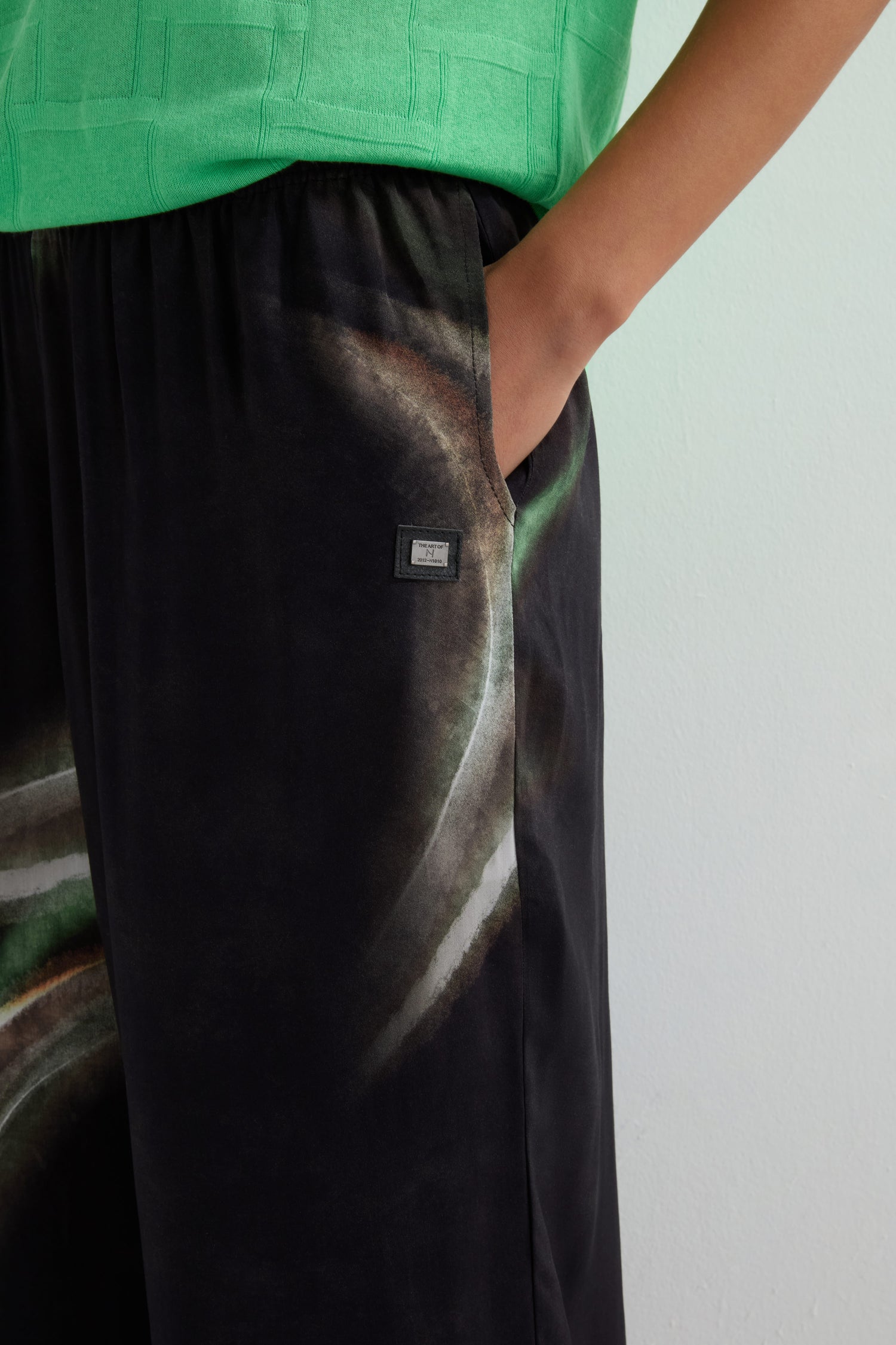 A person wearing a green top and Aurora Print Straight Trouser with a wide-leg silhouette stands with one hand in their pocket.