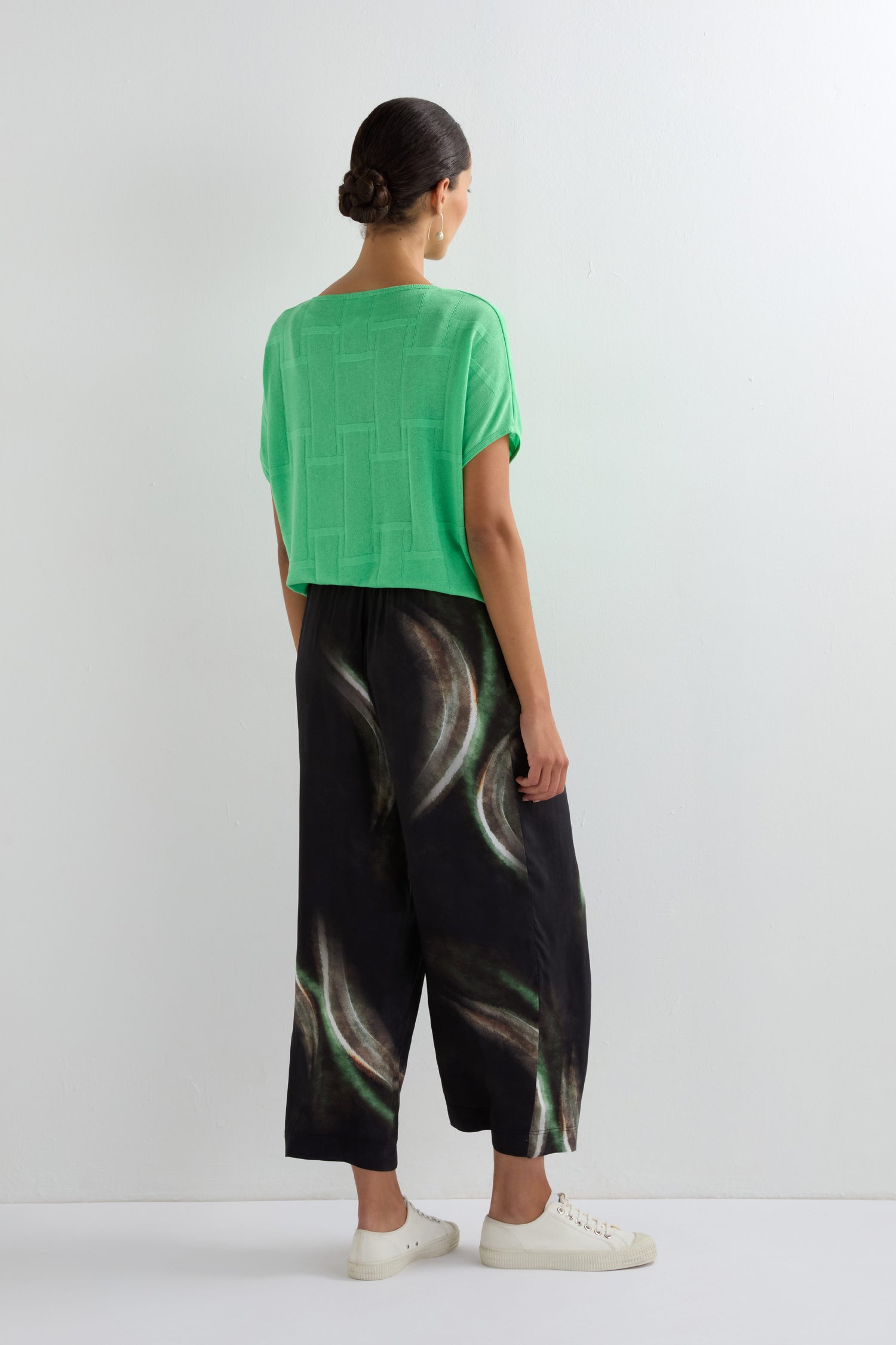 A person stands facing a white wall, wearing a bright green short-sleeve top, Aurora Print Straight Trouser with an elasticated waistband and wide-leg silhouette, and white sneakers.