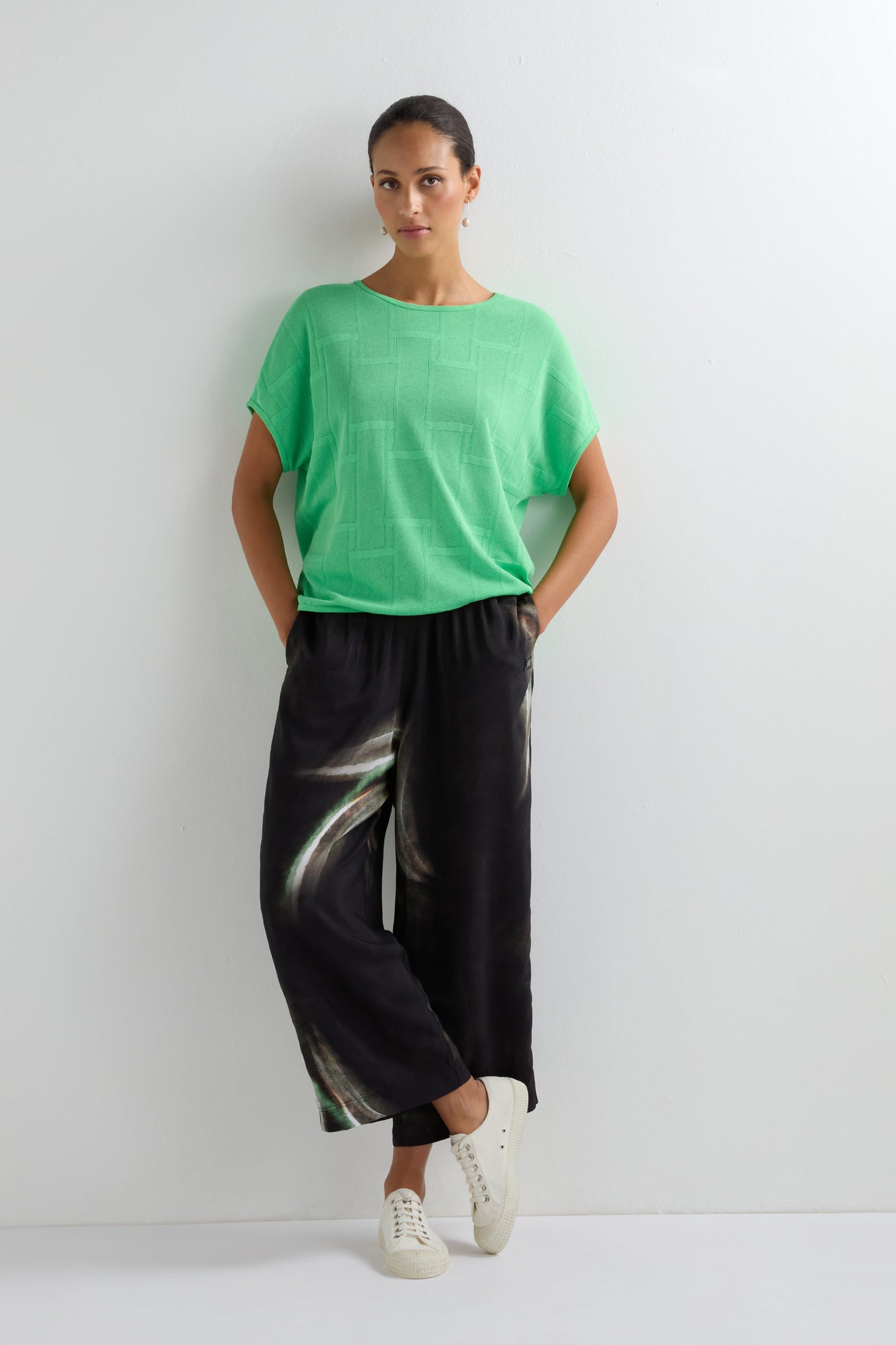 A woman stands against a white wall wearing a loose green top, Aurora Print Straight Trousers, and white sneakers, hands in her pockets.