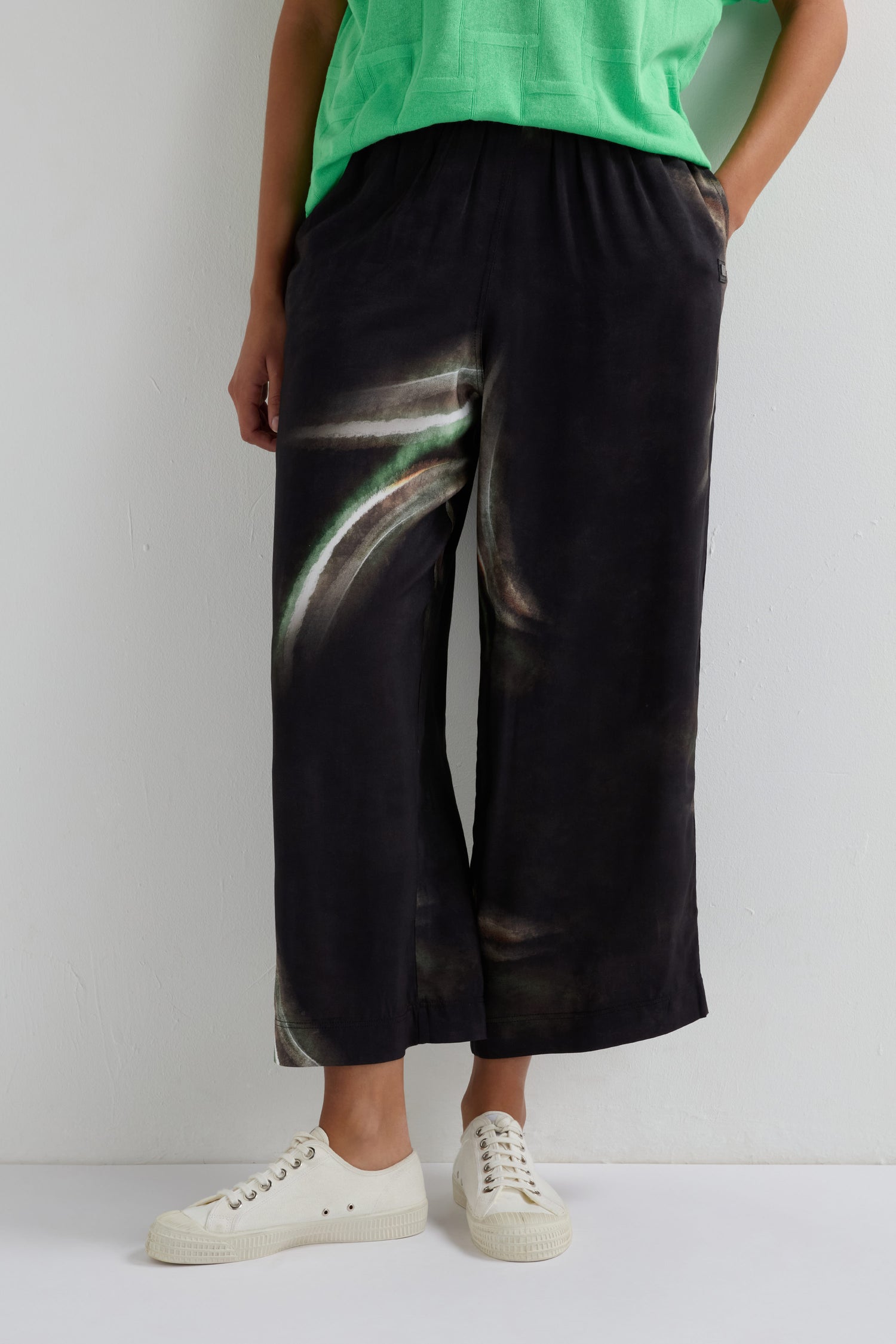 A person wears a green top, Aurora Print Straight Trouser featuring an elasticated waistband and wide-leg silhouette, paired with white sneakers, standing against a white wall.
