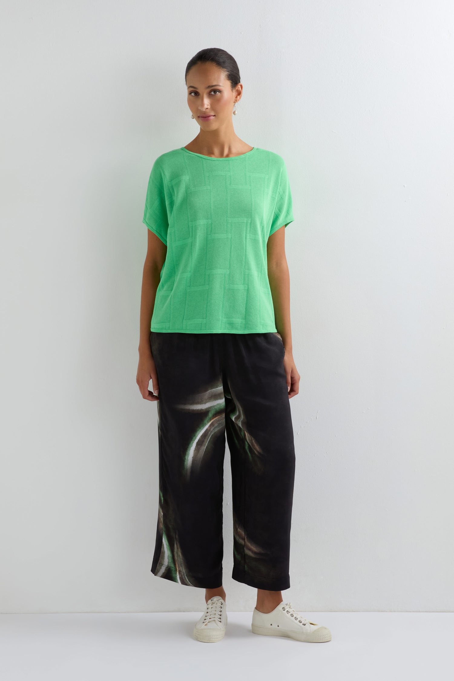 A woman stands against a white wall, wearing the Grid Texture Boxy Knit in light green, wide-leg black pants with a wavy pattern, and white sneakers.
