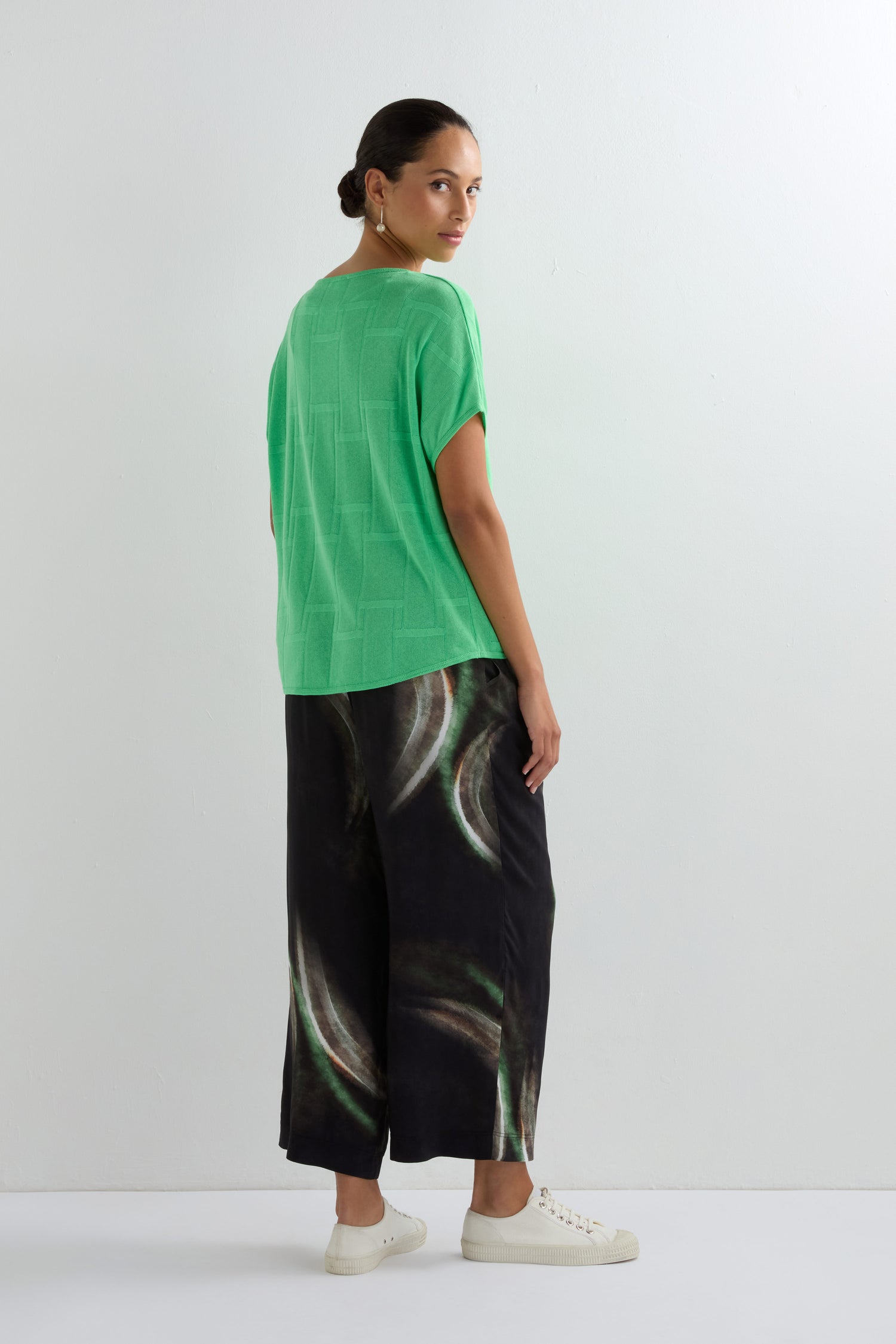 A woman stands sideways against a white background wearing the Grid Texture Boxy Knit in green, wide-leg black pants with a circular pattern, and white sneakers.