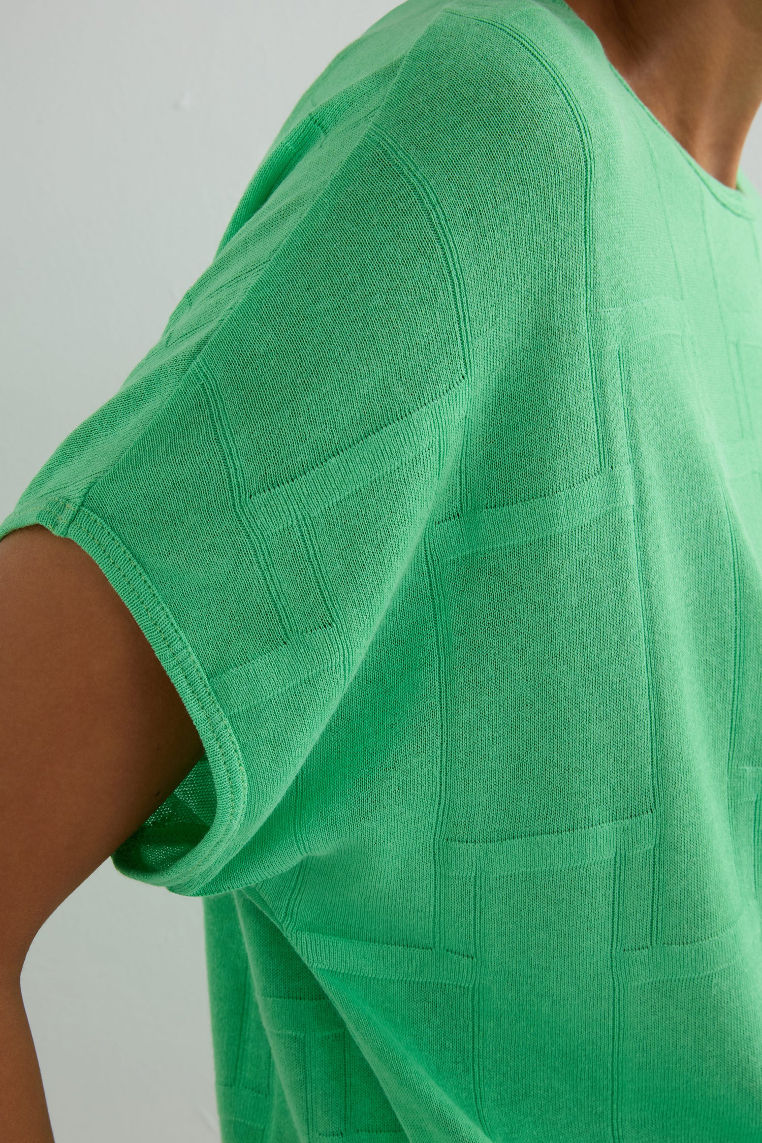 Close-up of a person wearing the Grid Texture Boxy Knit, a green short-sleeve top with a subtle square pattern, against a light background.