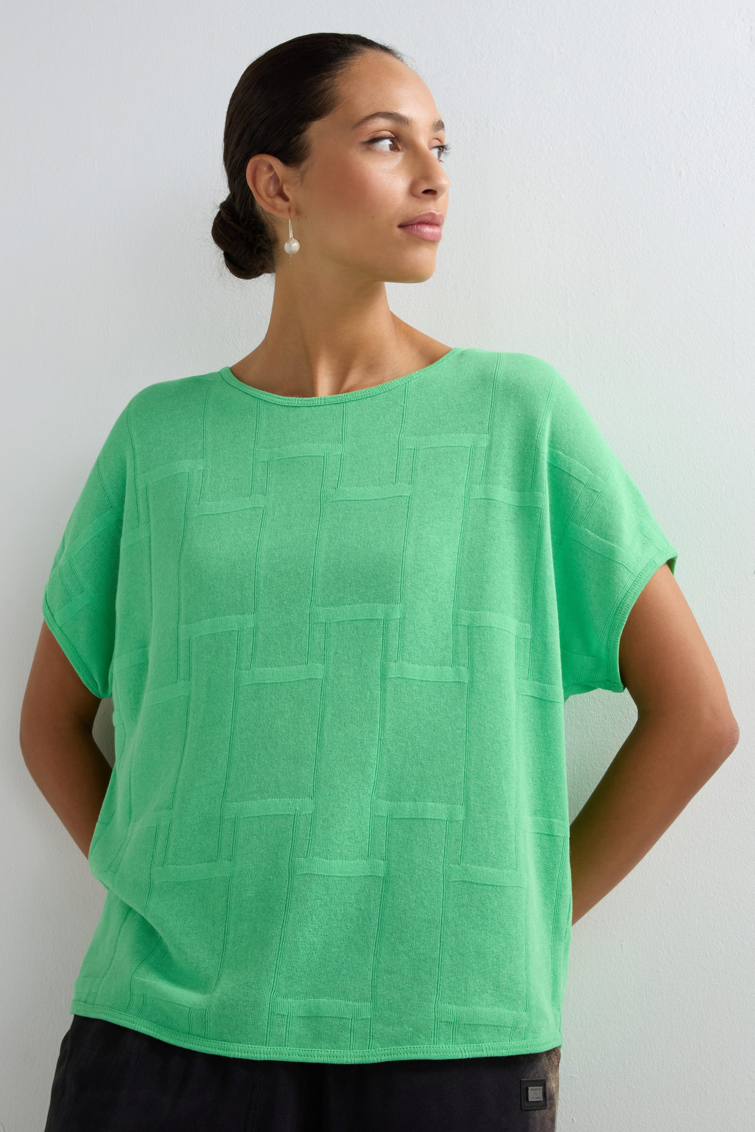 A woman stands against a white wall, looking sideways. She wears the Grid Texture Boxy Knit in light green with pearl drop earrings, and her hair is styled back in a neat bun.