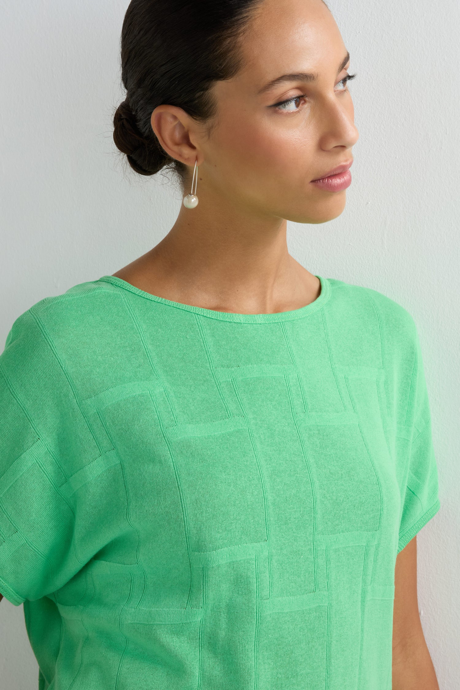 A woman with dark hair in a low bun wears the Grid Texture Boxy Knit in light green and a single dangling earring, gazing to the side against a plain background.