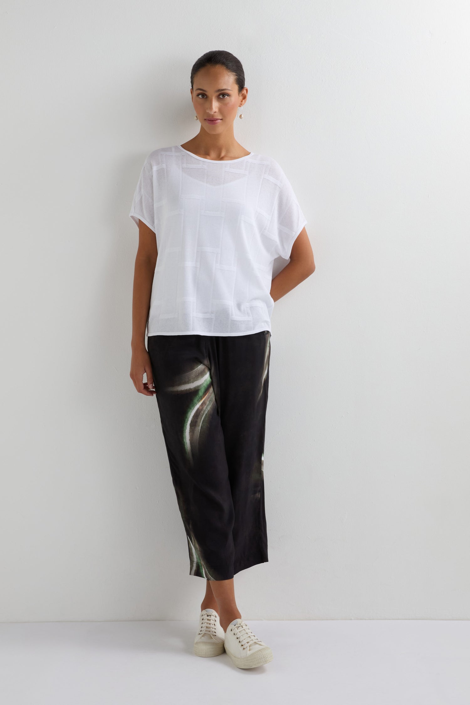 A person stands against a white wall wearing the Grid Texture Boxy Knit white short-sleeve top, black wide-leg pants with a green abstract pattern, and white sneakers.