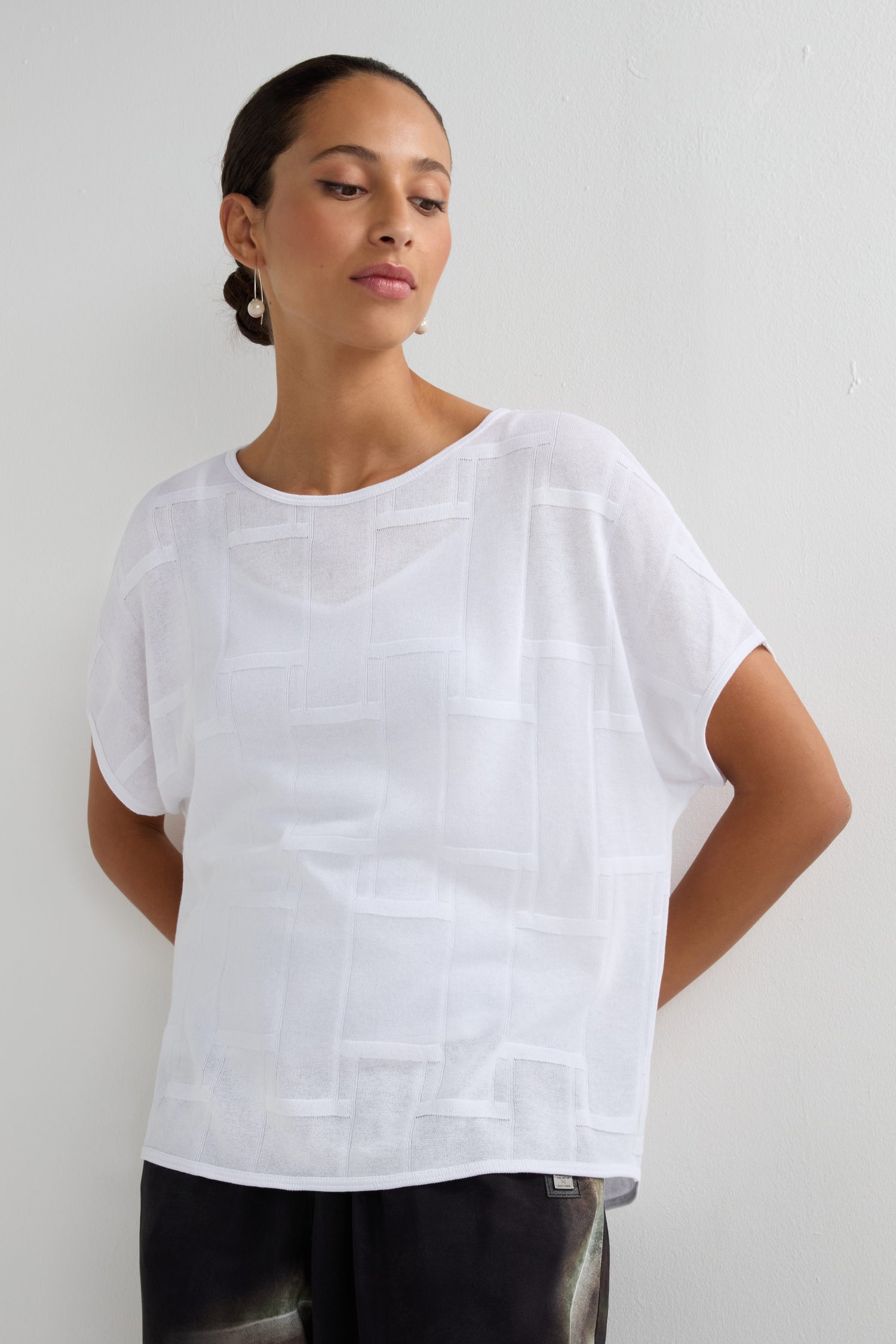 A woman stands against a light wall wearing the Grid Texture Boxy Knit, a loose-fitting white mesh short-sleeve top, paired with dark pants and her hands behind her back.