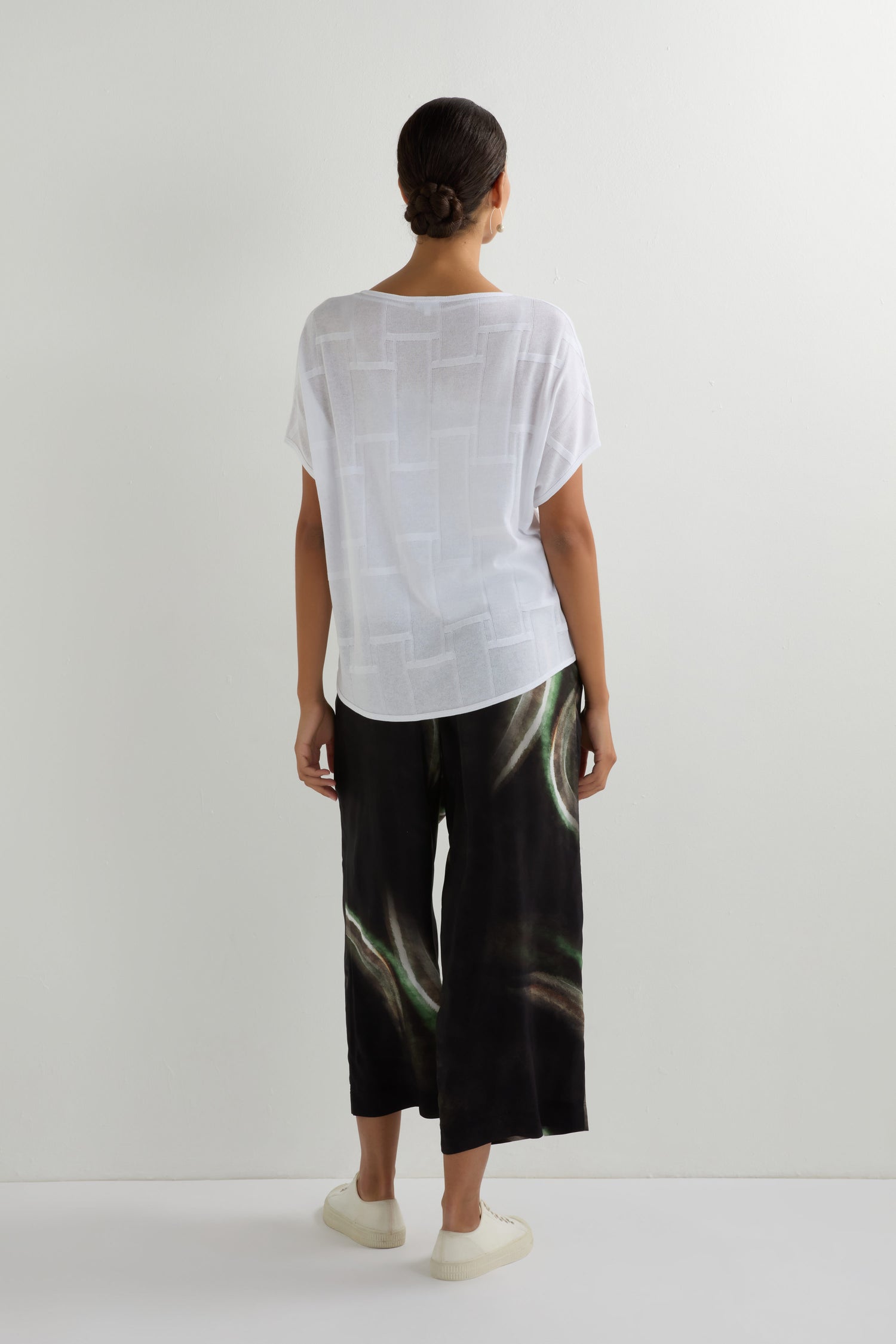 A person stands facing a white wall, wearing the Grid Texture Boxy Knit in sheer white short sleeves, wide-leg black pants with abstract green patterns, and light-colored slip-on shoes.