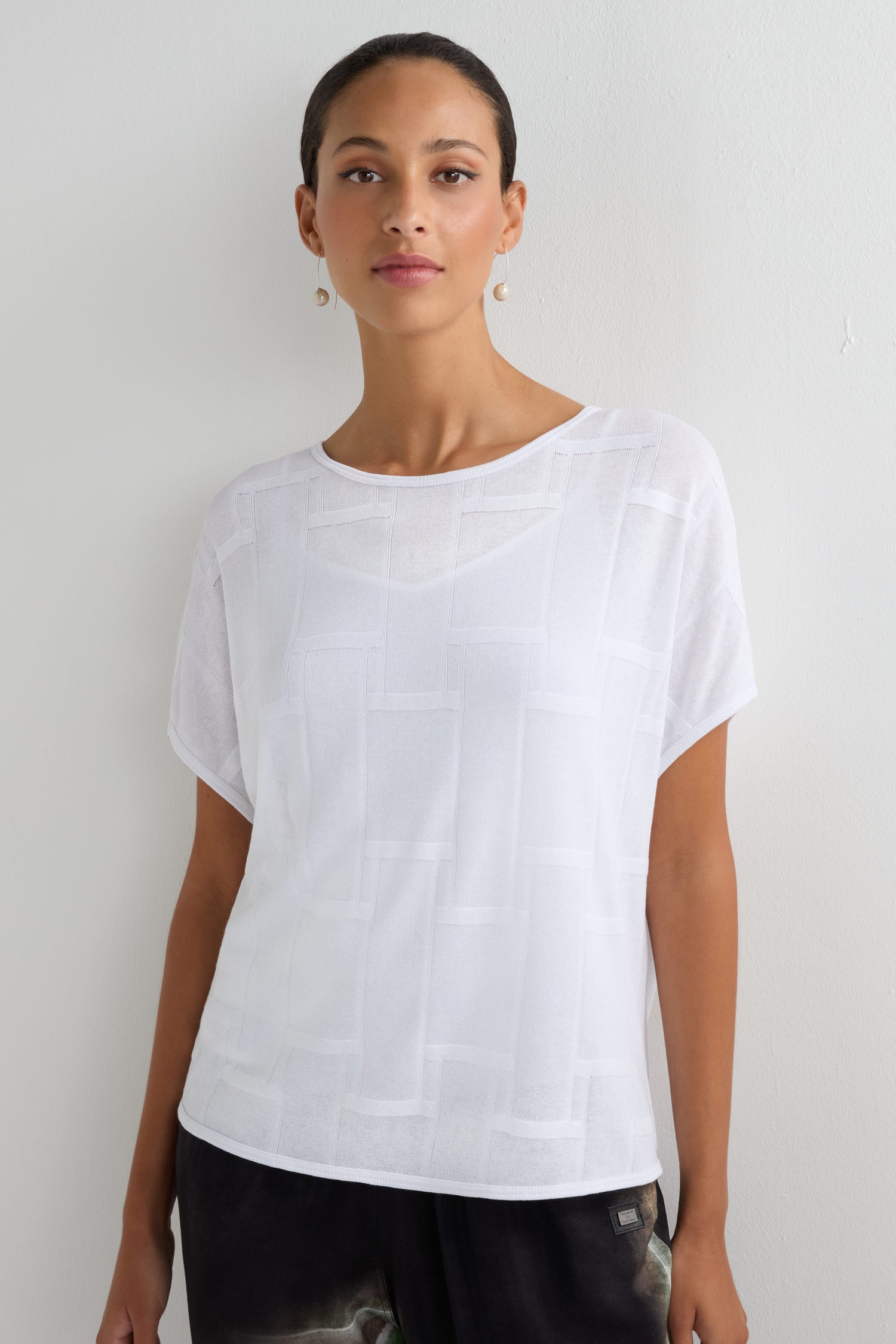 A woman stands against a white wall wearing the Grid Texture Boxy Knit, a short-sleeved white textured top, paired with dark pants, her hair pulled back and accessorized with pearl drop earrings.