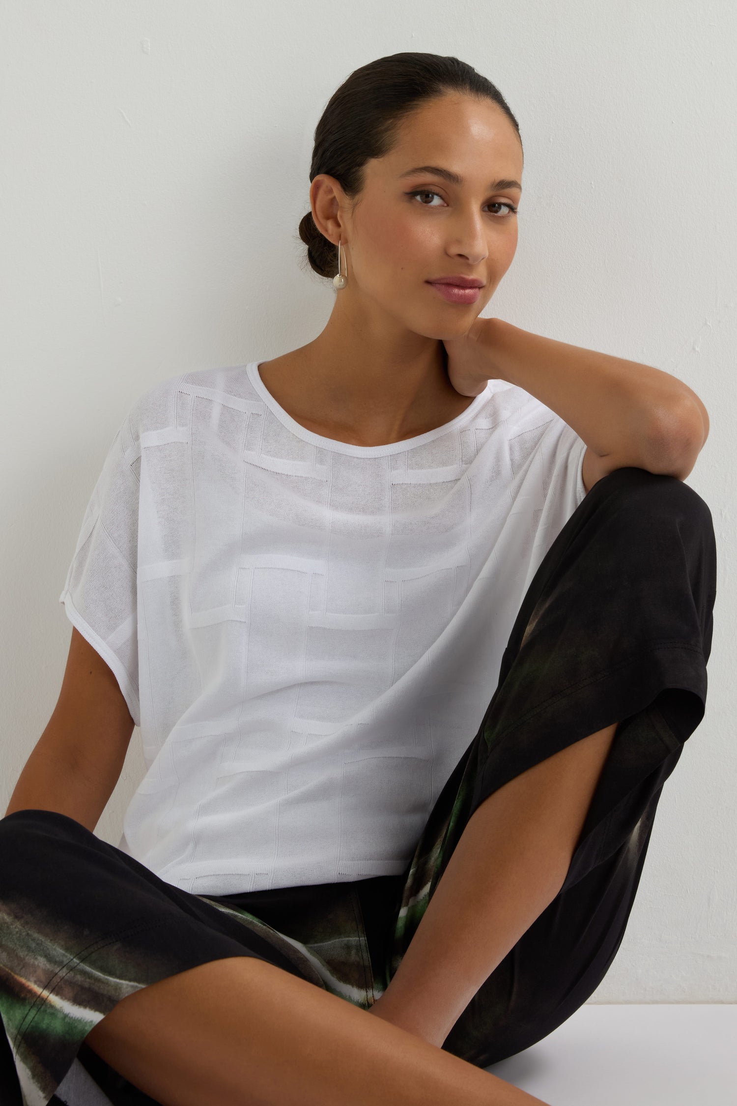 A woman with tied-back hair sits against a white wall, wearing the Grid Texture Boxy Knit in sheer white and black pants with green patterns, looking at the camera with a relaxed expression.