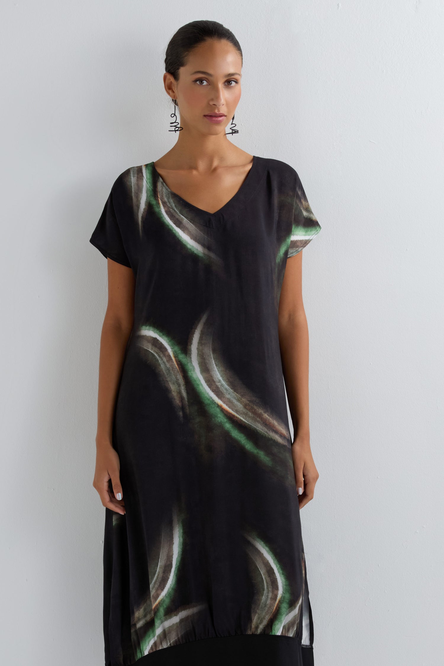 Woman with dark hair tied back wears the Aurora Print V Neck Dress, a midi-length piece featuring green and white curved patterns, short sleeves, and a flattering V neckline. She completes her look with dangling earrings against a white wall.