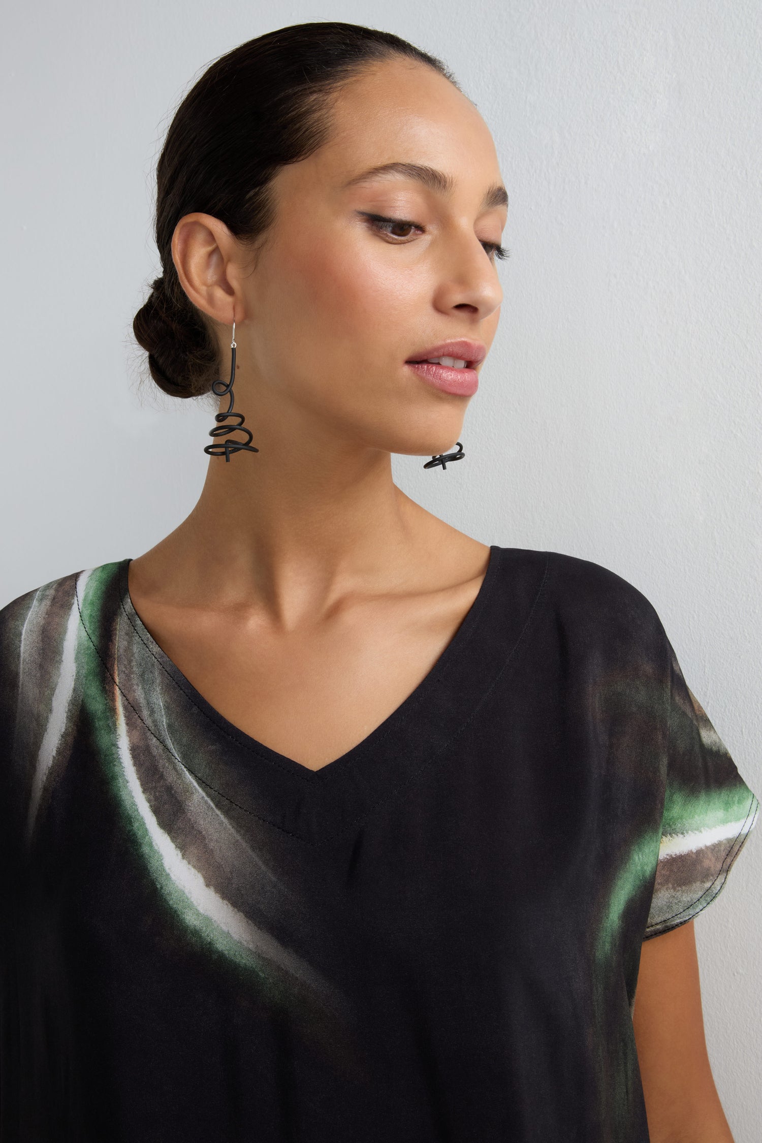 A woman with dark hair in a low bun wears the Aurora Print V Neck Dress, a black dress with green and white patterns, and large spiral earrings, standing against a plain light wall.