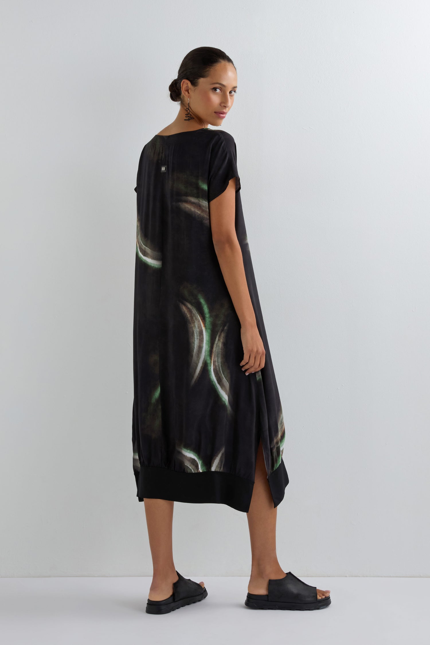A person stands sideways against a plain white wall wearing the Aurora Print V Neck Dress, which features a loose fit, v-neckline, black tie-dye pattern, and is styled with black slide sandals.