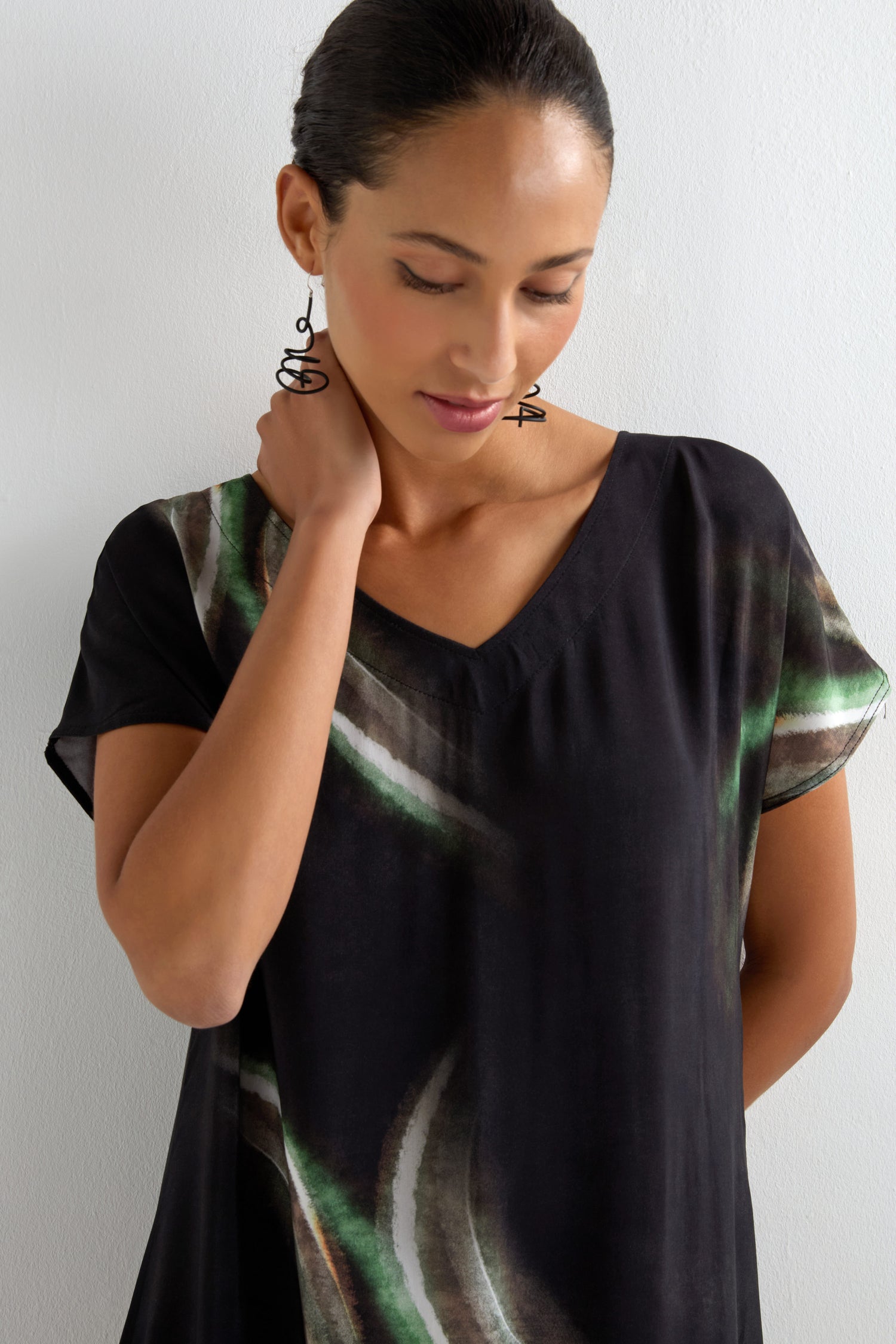 A woman with dark hair pulled back touches her neck, wearing the Aurora Print V Neck Dress—a black and green midi dress with a V-neck—plus spiral earrings, standing against a plain white wall.