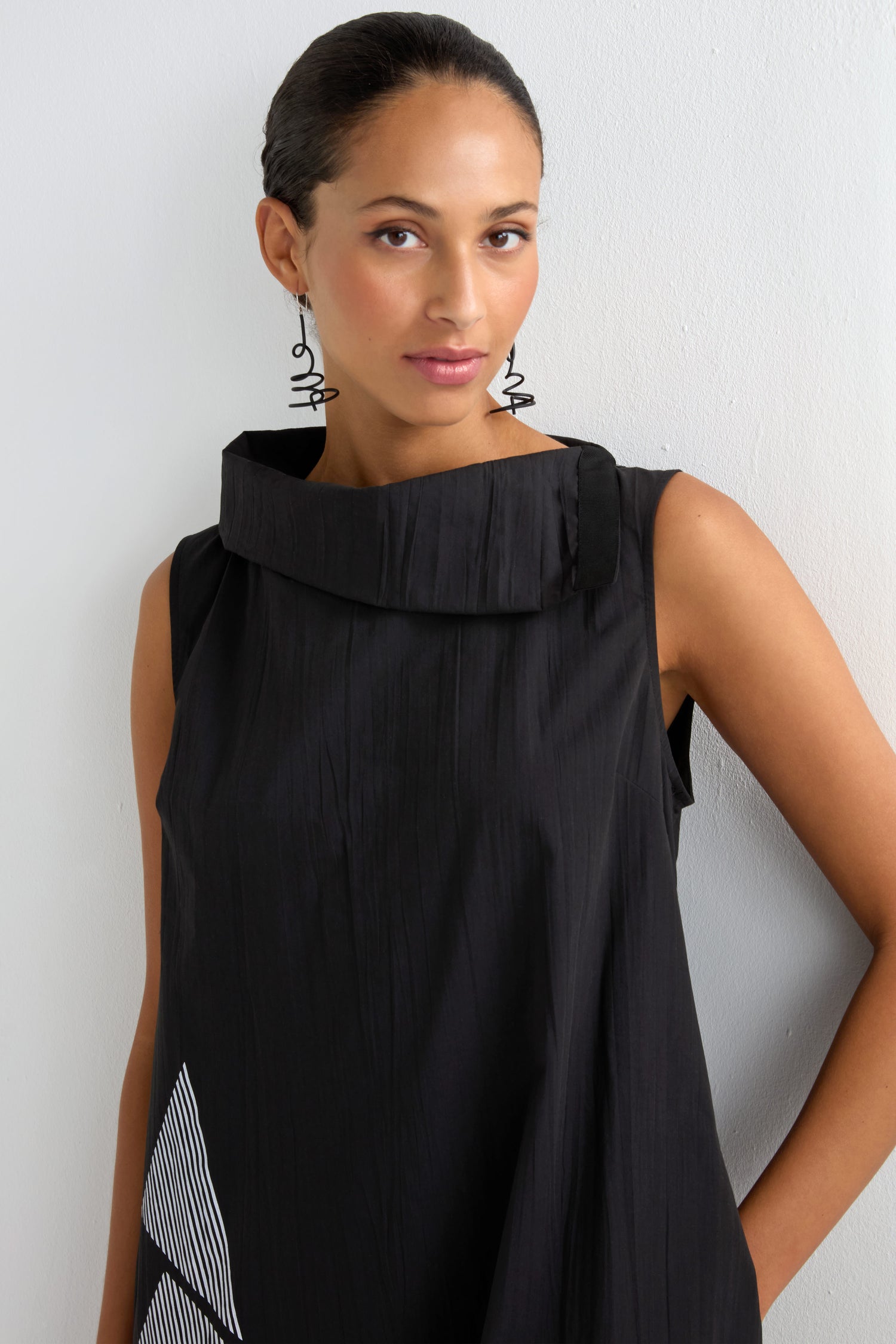 A woman with tied-back hair wears a sleeveless black Roll Neck Placement Print Crinkle Dress and geometric earrings, standing against a plain white wall.