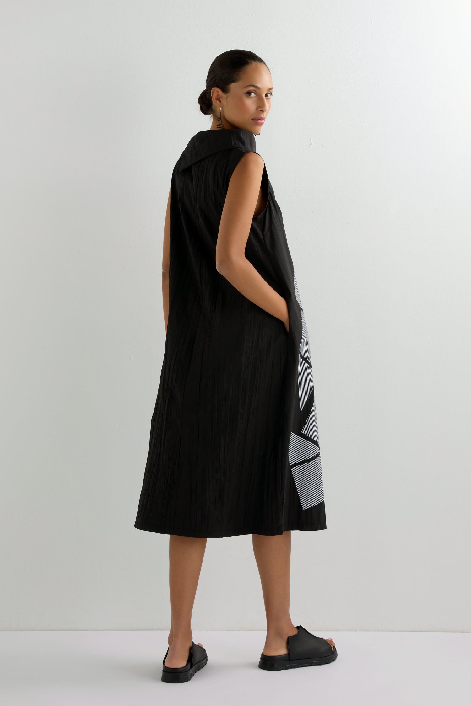 A woman in a sleeveless black Roll Neck Placement Print Crinkle Dress with bold side patterns stands turned slightly away, glancing over her shoulder.