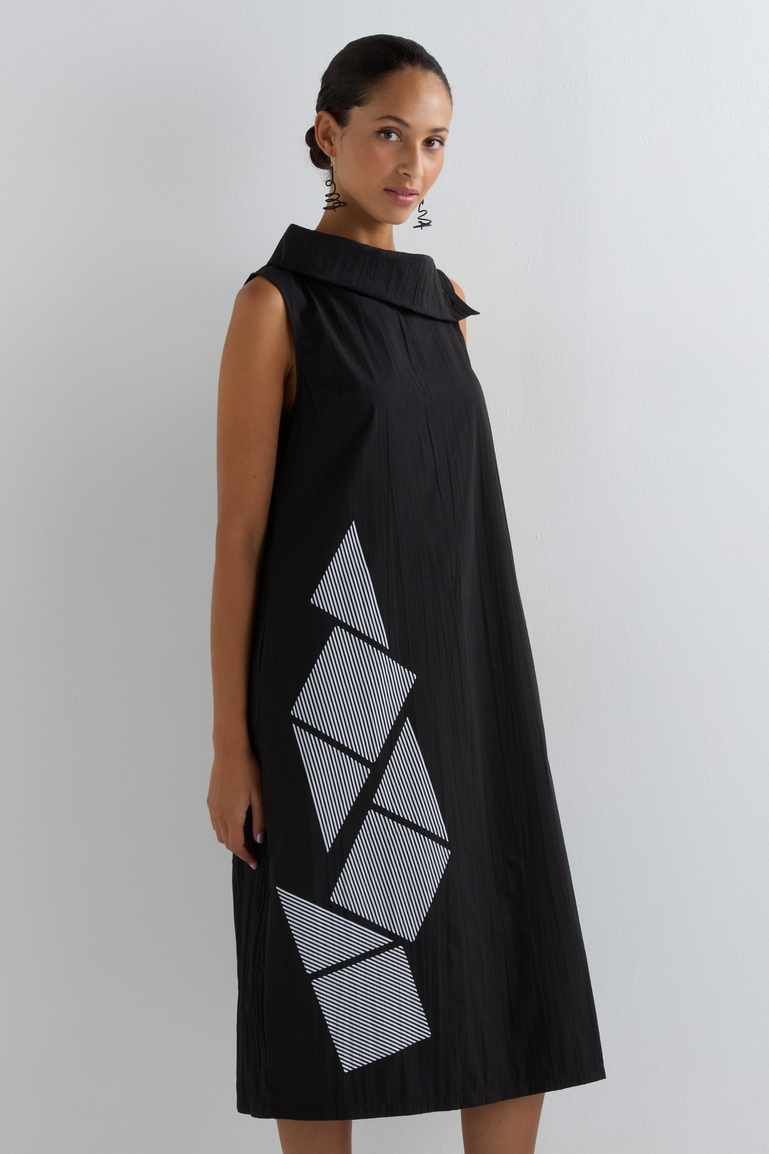 A woman wearing the Roll Neck Placement Print Crinkle Dress, a sleeveless black dress with a high collar and bold white and gray geometric patterns, stands against a plain light background.