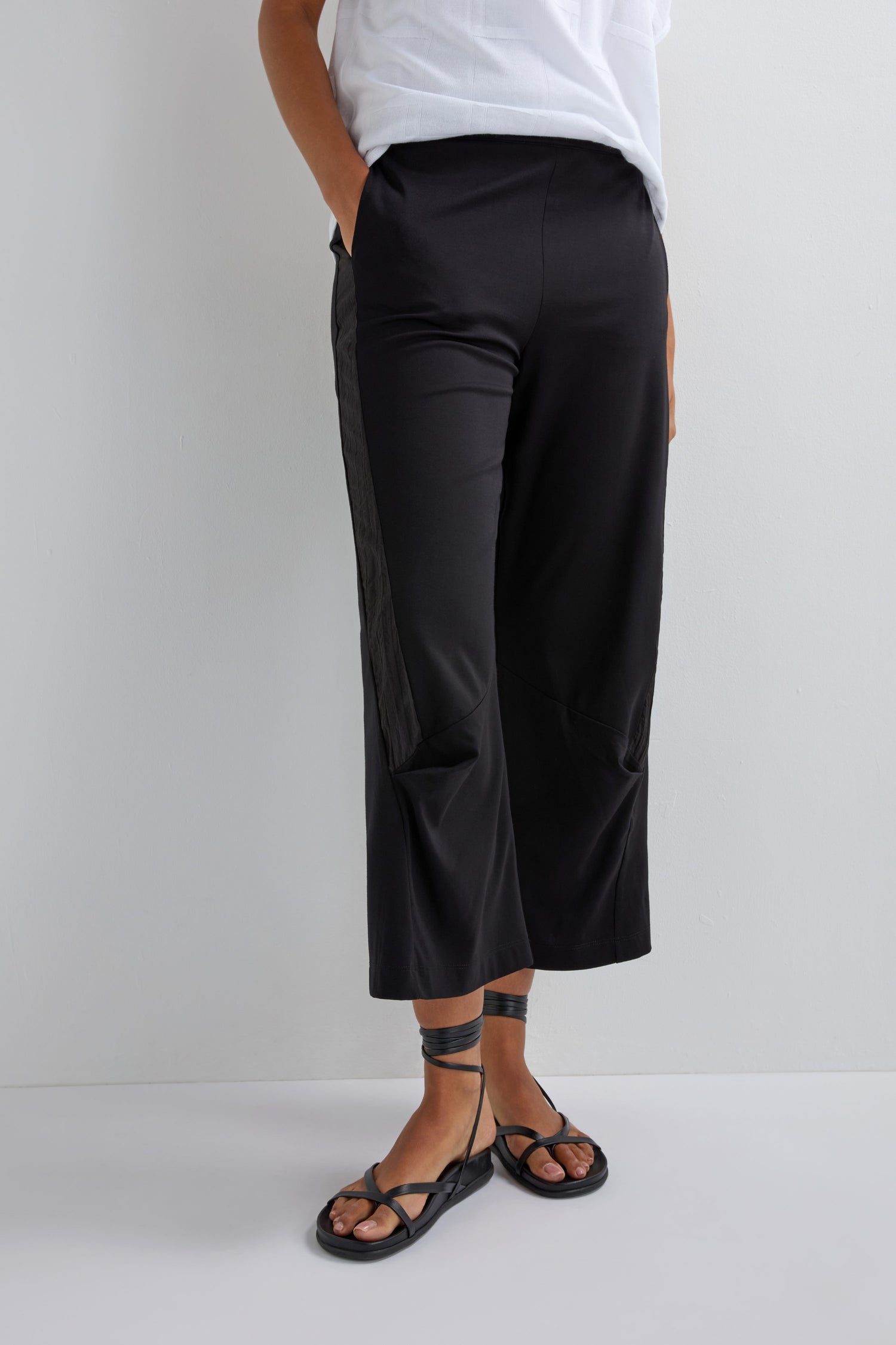 A person stands against a plain white wall in Crinkled Panel Trouser, featuring stretch fabric and side detailing, paired with a white top and black strappy sandals for a modern look.