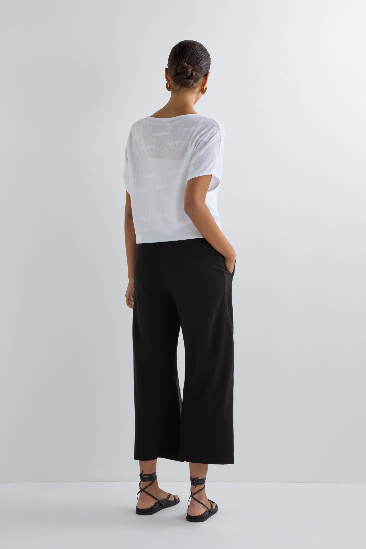 A person stands facing away against a plain white wall, wearing a sheer white t-shirt, Crinkled Panel Trouser for a modern look, and black strappy sandals with hands in their pockets.