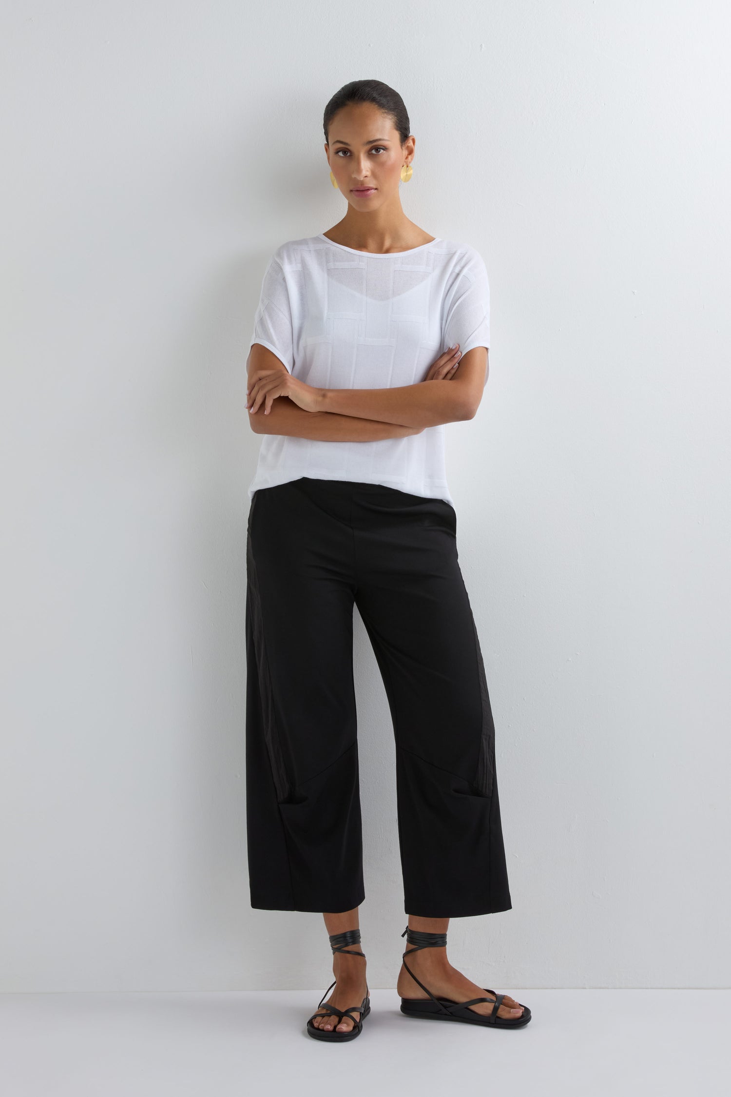 A person stands against a white wall, wearing a white short-sleeve top, black Crinkled Panel Trousers, and black sandals, arms crossed and looking at the camera for a modern look.