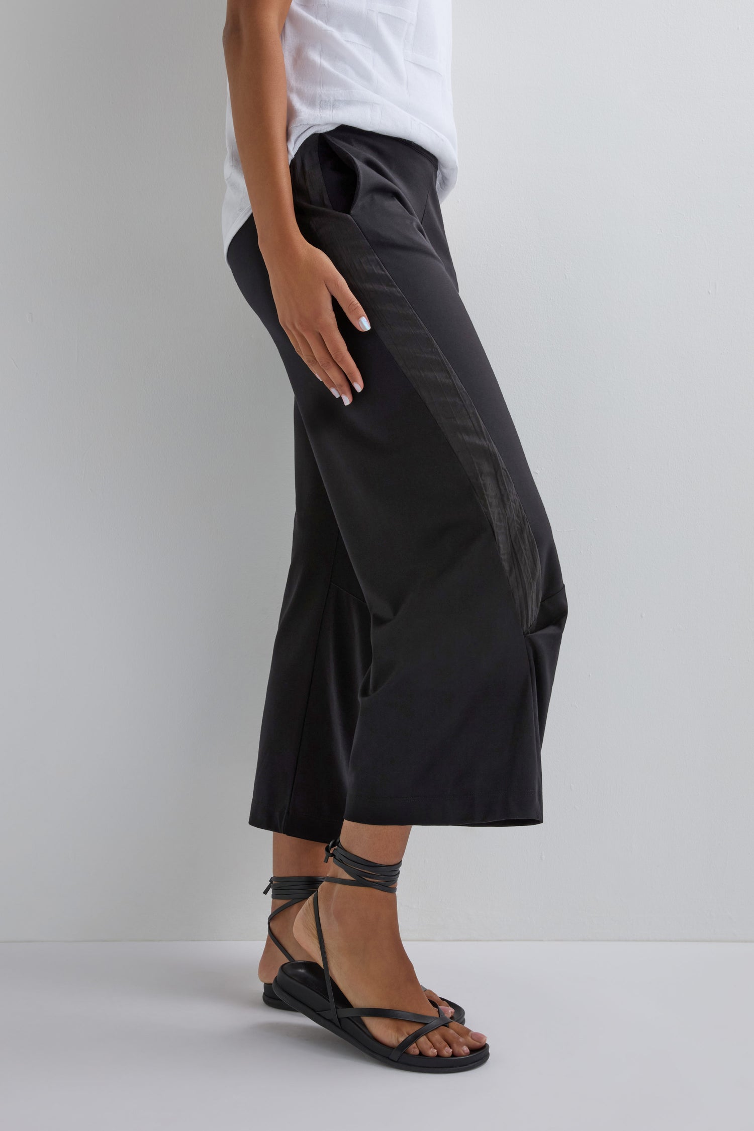 Wearing the Crinkled Panel Trouser in black with a white shirt and black strappy sandals, the person stands against a plain white background, showcasing a contemporary-edge look.