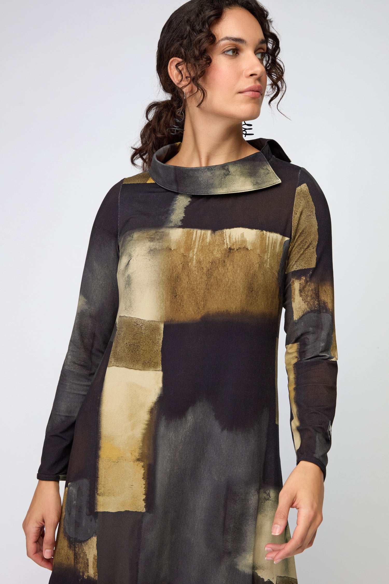 A woman stands looking to the side, wearing the Block Print Collar Jersey Dress featuring long sleeves and a black and gold rectangular abstract pattern.