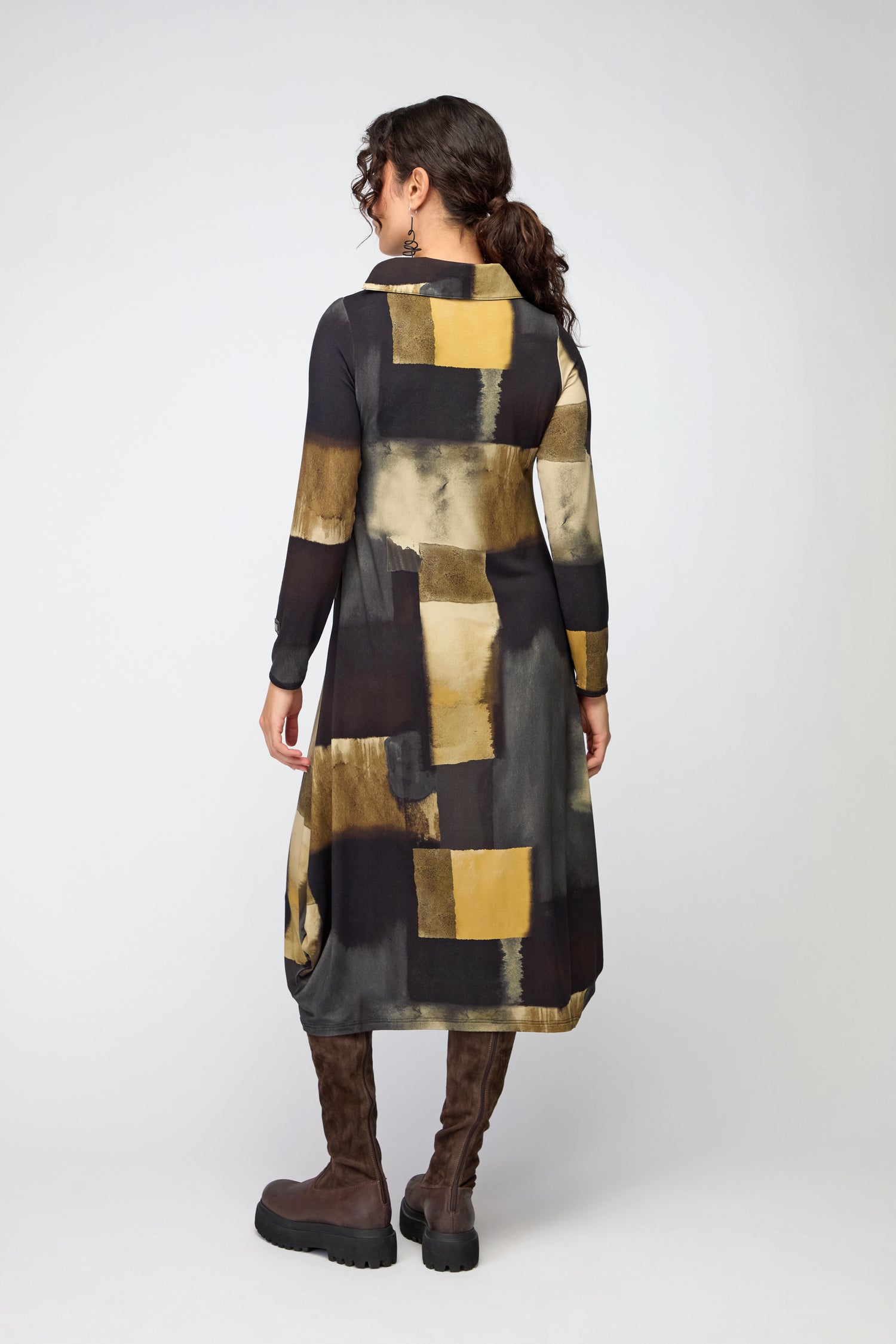 A woman faces away, wearing a yellow and black Block Print Collar Jersey Dress paired with brown knee-high boots and earrings against a plain background.