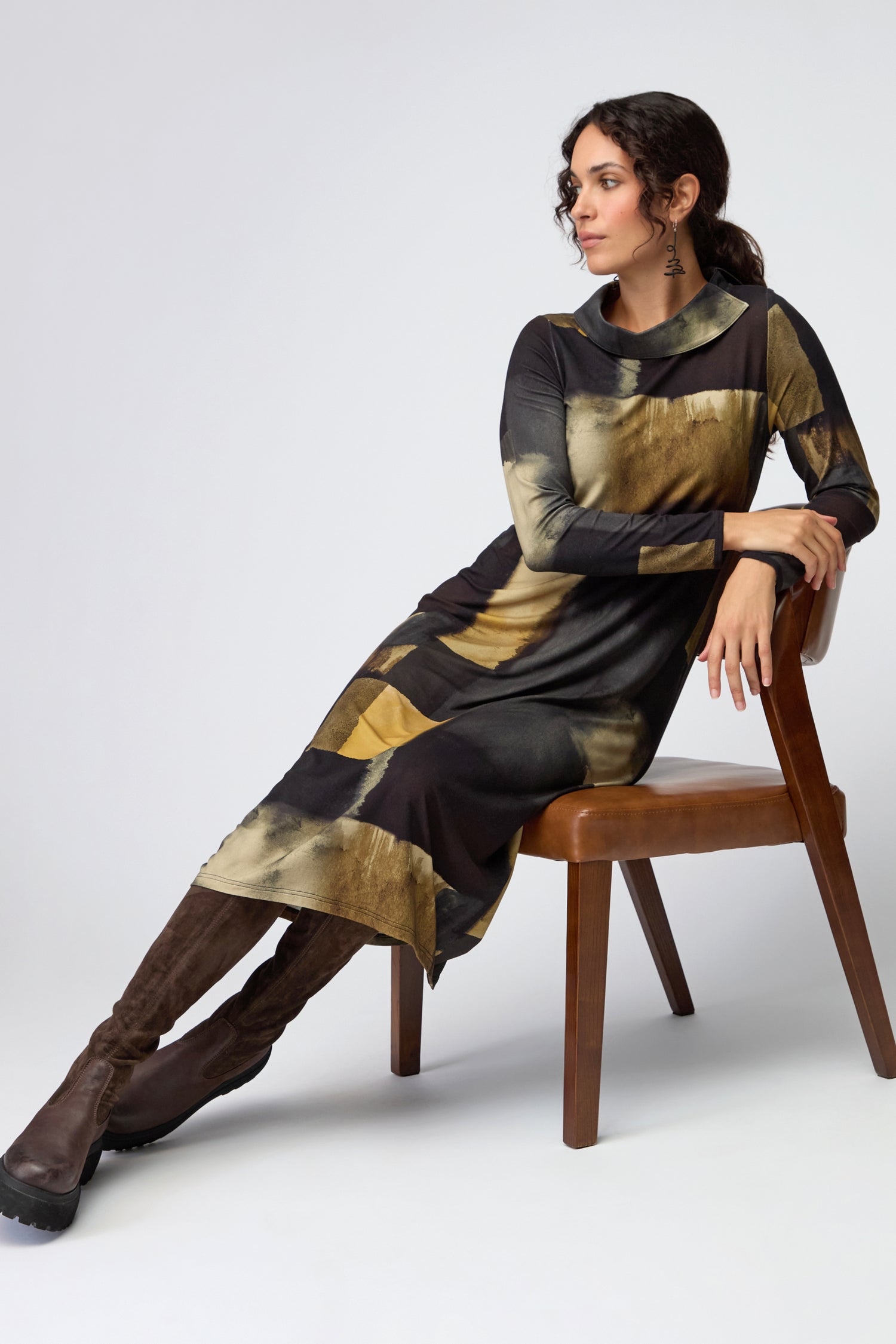 A woman in the Block Print Collar Jersey Dress, featuring long sleeves and a black and gold pattern, sits sideways on a wooden chair with one arm resting on the back and wears brown knee-high boots.
