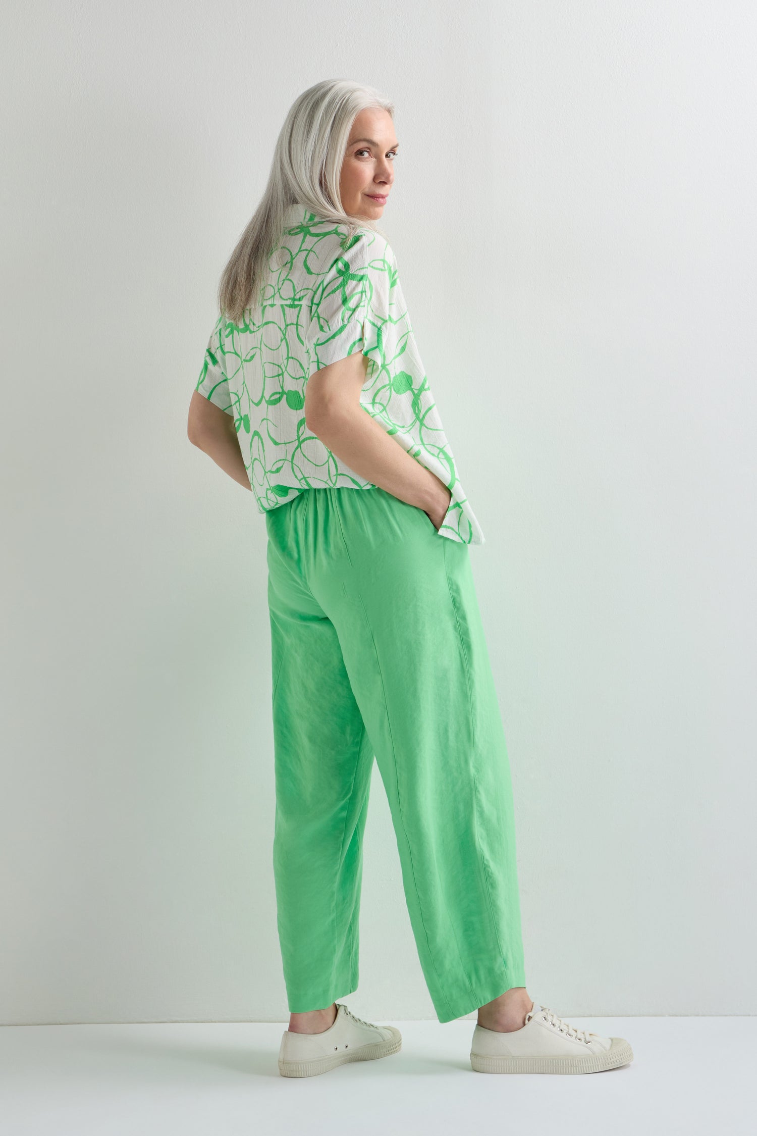 A woman with long gray hair stands sideways against a white background, wearing the Leaf Green Bubble Trouser in a lightweight cotton blend and a sculptural edge patterned shirt, paired with white sneakers.