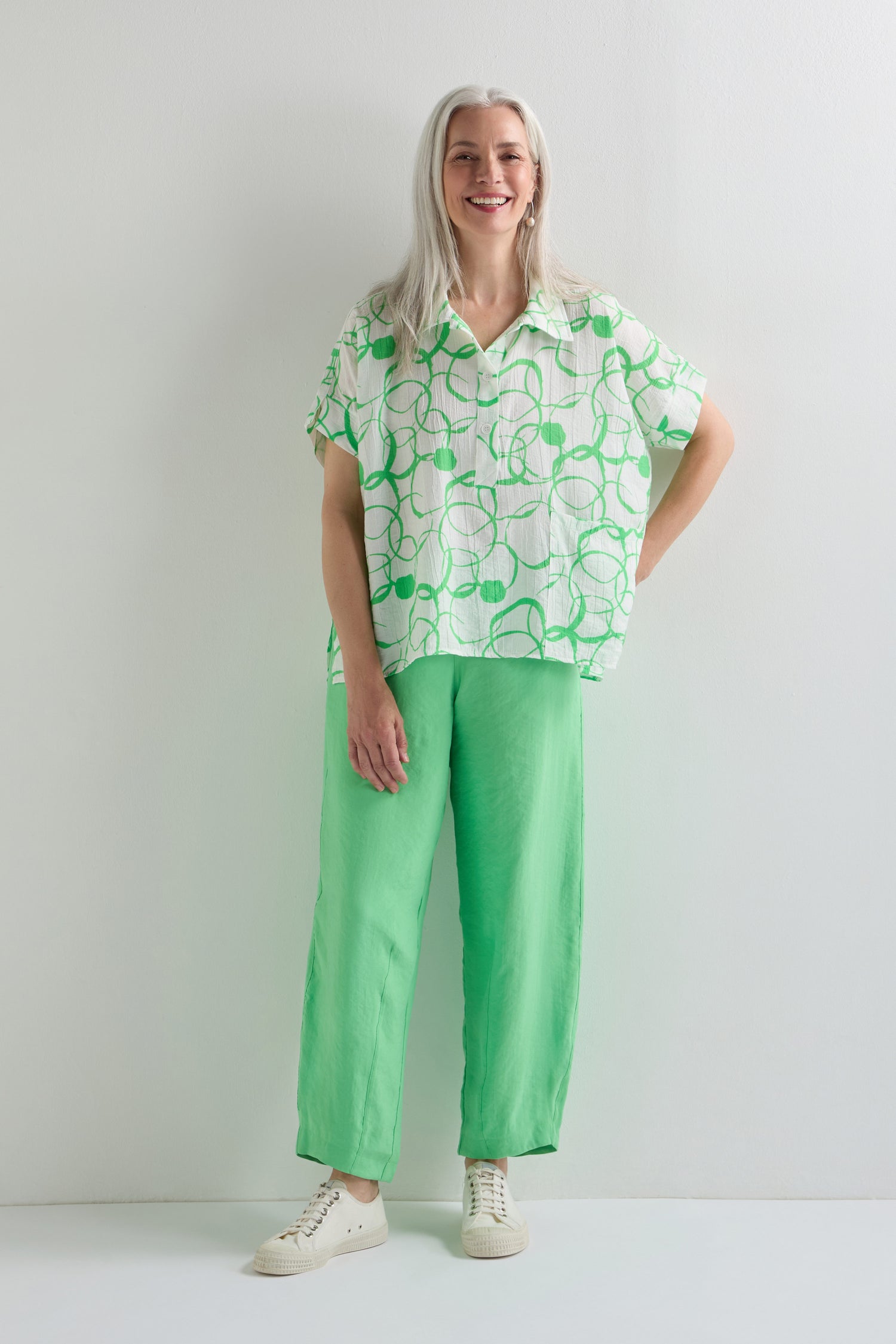 A woman with long gray hair wears leaf green bubble trousers, a lightweight cotton blend white shirt with a green abstract pattern, and white sneakers, standing against a plain white background.