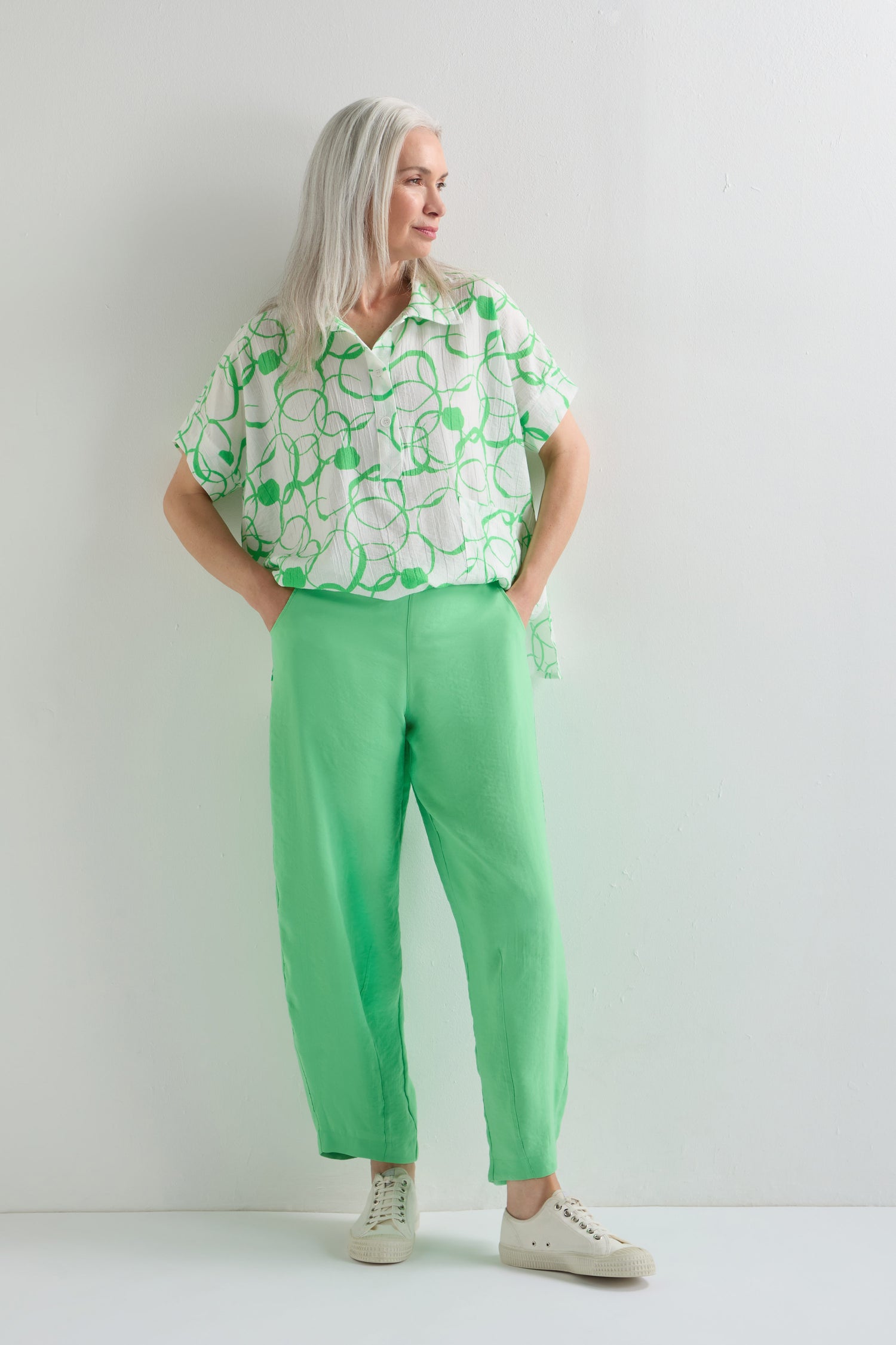 A woman with long gray hair stands against a white wall, wearing a patterned short-sleeve shirt, the Leaf Green Bubble Trouser in a lightweight cotton blend, and white sneakers.