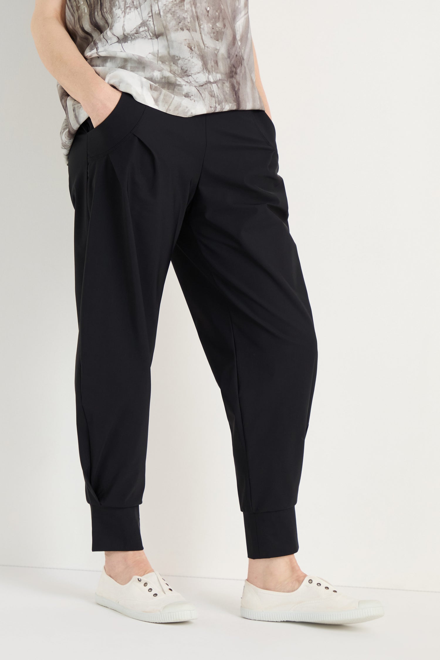 Person stands with hands in pockets, wearing Tapered Cuff Trousers made from stretch fabric, a patterned top, and white slip-on sneakers against a plain background.