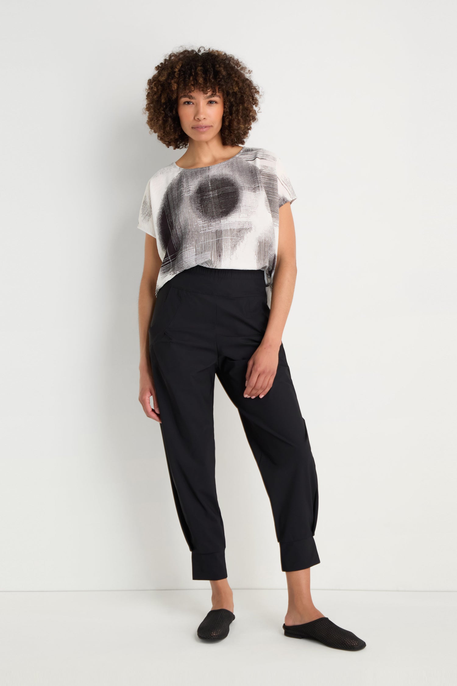 Person stands with hands in pockets, wearing Tapered Cuff Trousers made from stretch fabric, a patterned top, and white slip-on sneakers against a plain background.