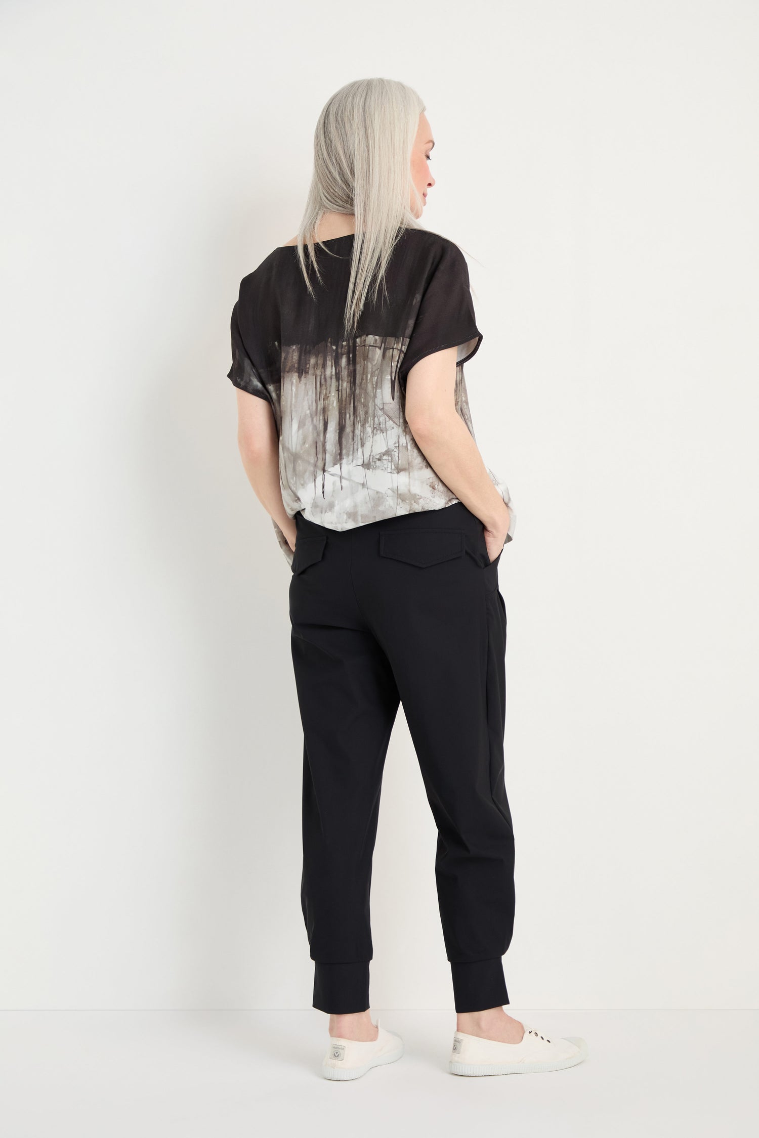 A person with long gray hair stands facing away, wearing a black and white short-sleeve top, Tapered Cuff Trousers, and white sneakers against a plain white background.