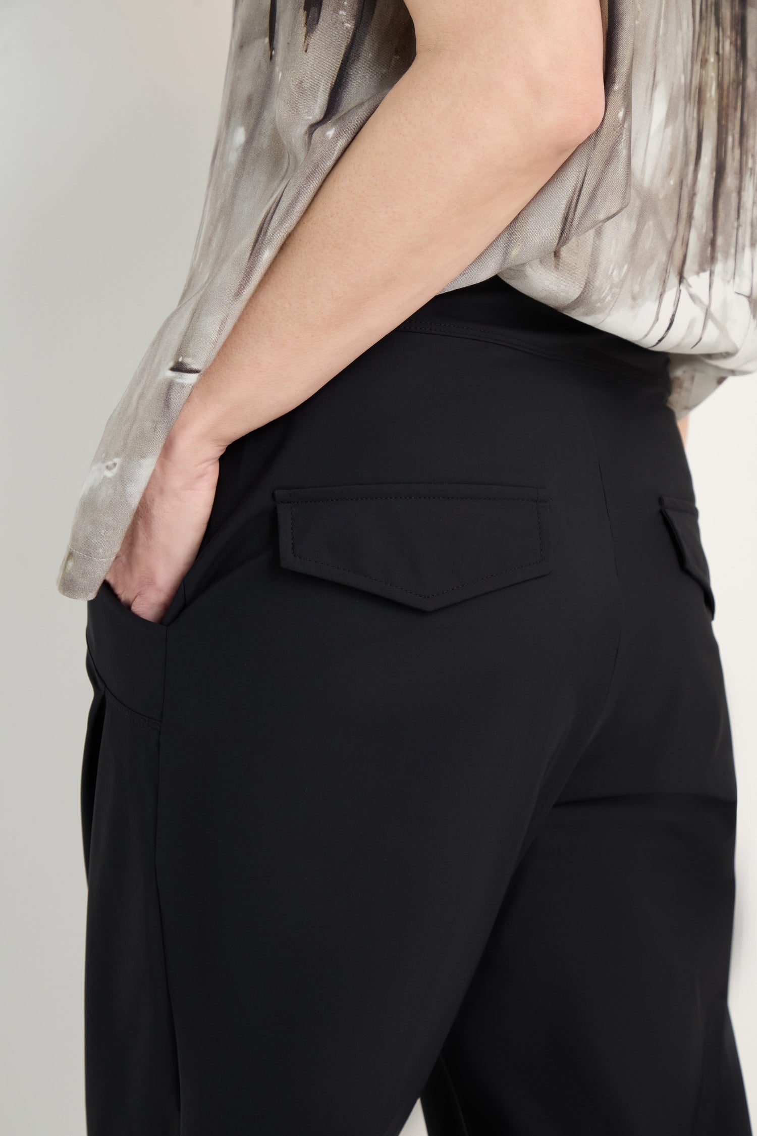 A person wearing black Tapered Cuff Trousers with a flap pocket has their left hand in the pocket; only their lower torso and part of an abstract-patterned shirt are visible.