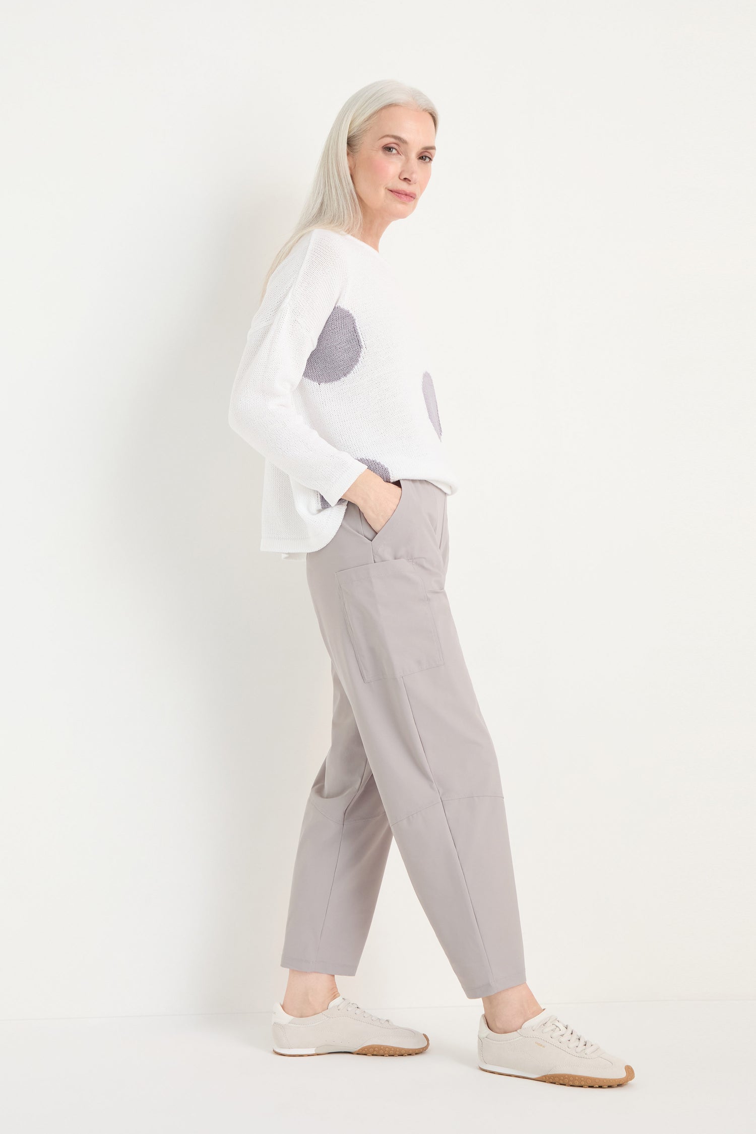 A woman with long gray hair wears a white sweater, cropped light gray Tapered Trousers in stretch fabric, and beige sneakers, posed against a plain white background.