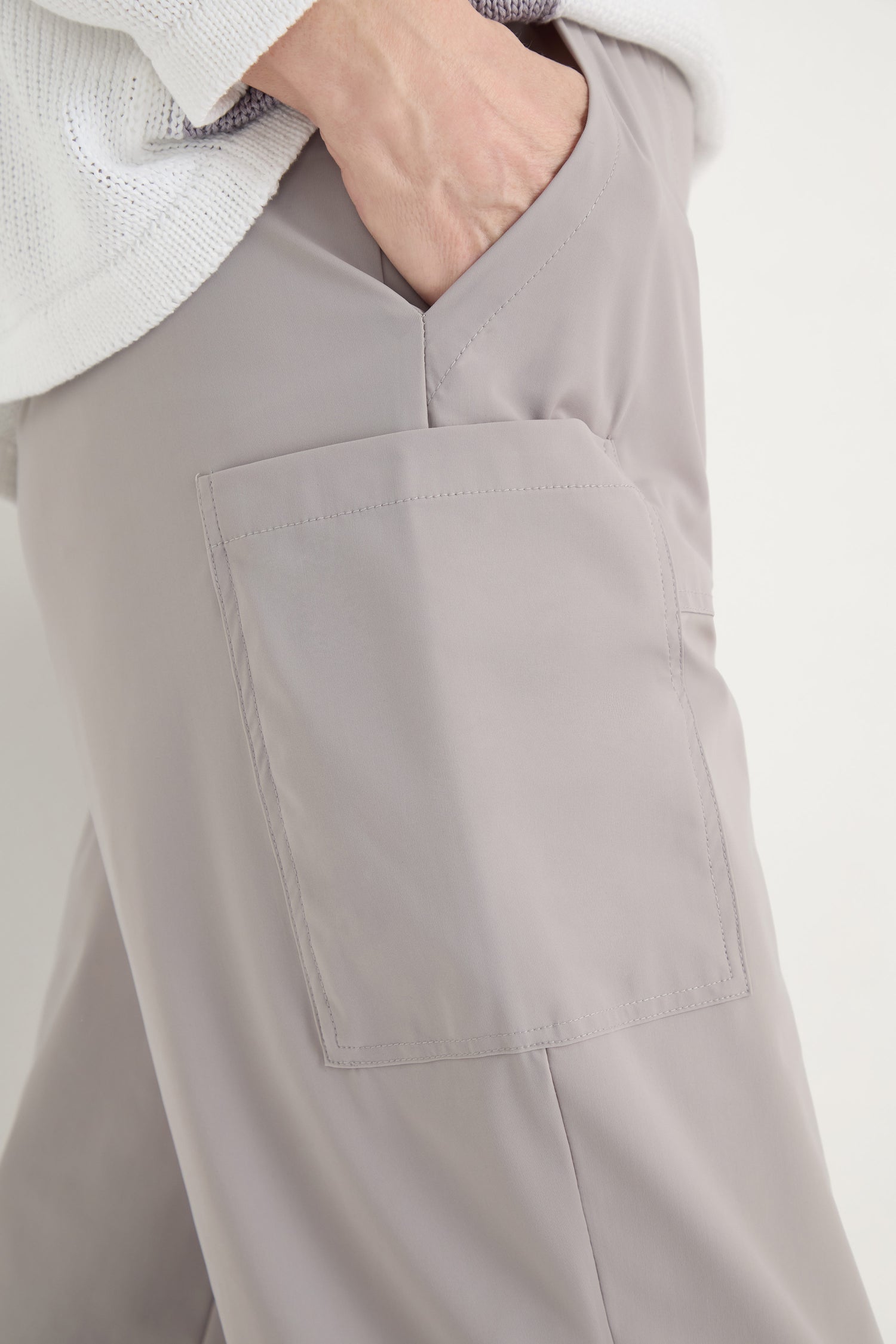 A person wears Tapered Trousers in light gray with a large front pocket and a cropped fit, one hand tucked in. They pair the trousers with a white textured top for a stylish look.