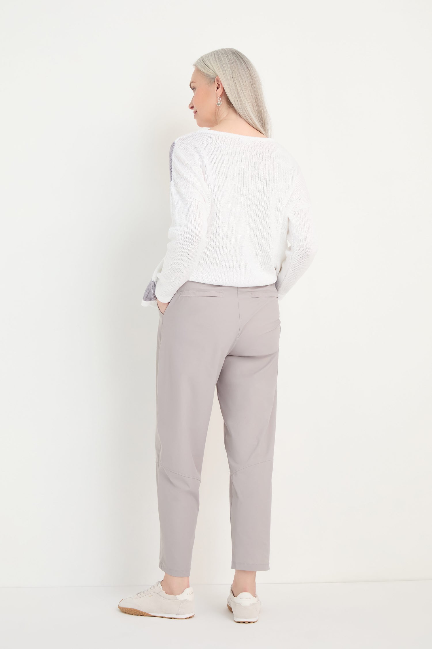 A person with long gray hair, seen from behind, wears a white sweater, Tapered Trousers in cropped stretch fabric, and white sneakers against a plain white background.